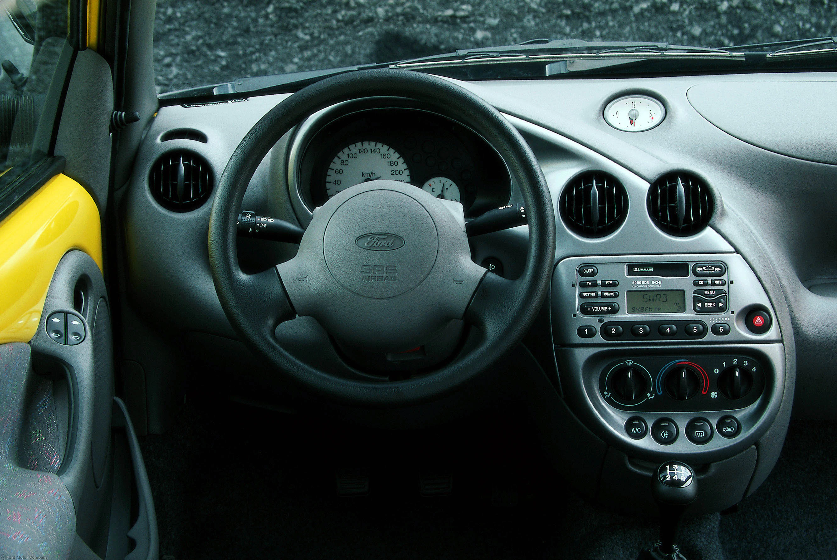 Innenraum eines Autos mit grauem Armaturenbrett, Lenkrad und integriertem Autoradio. Links ist ein Teil der gelben Fahrzeugtür sichtbar.