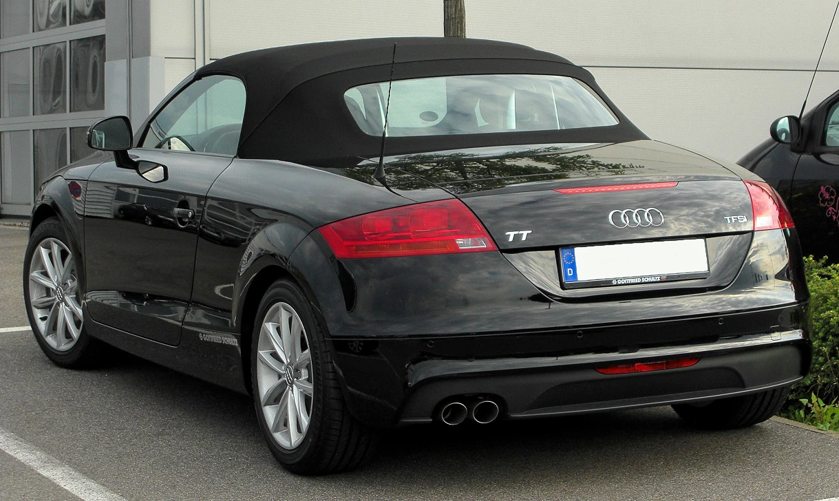 Ein schwarzer Audi TT Roadster mit geschlossenem Stoffverdeck, geparkt auf einem Parkplatz vor einem Gebäude.