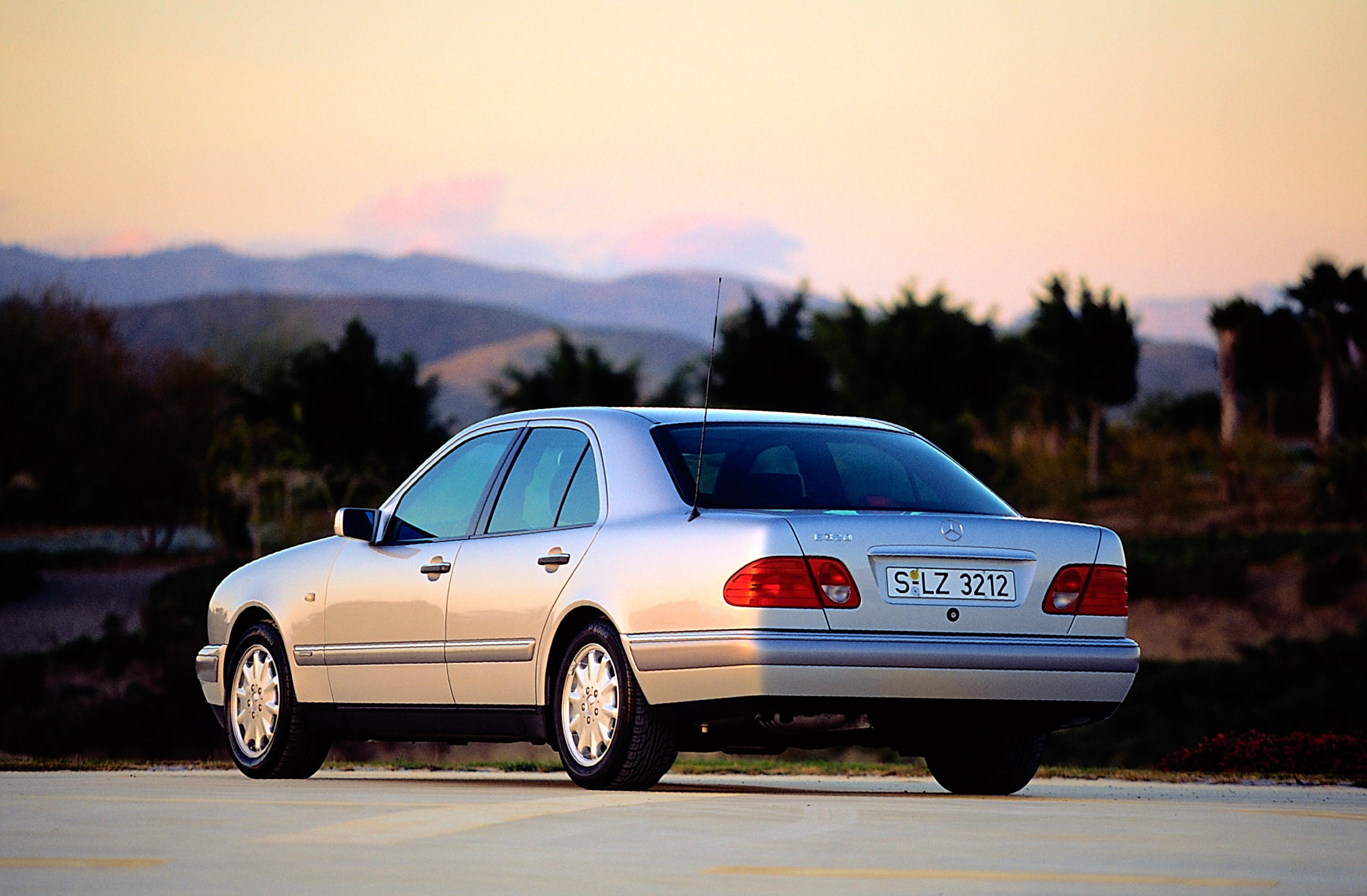 Silberner Mercedes-Benz Limousine geparkt im Freien bei Sonnenuntergang, mit Hügeln und Bäumen im Hintergrund.