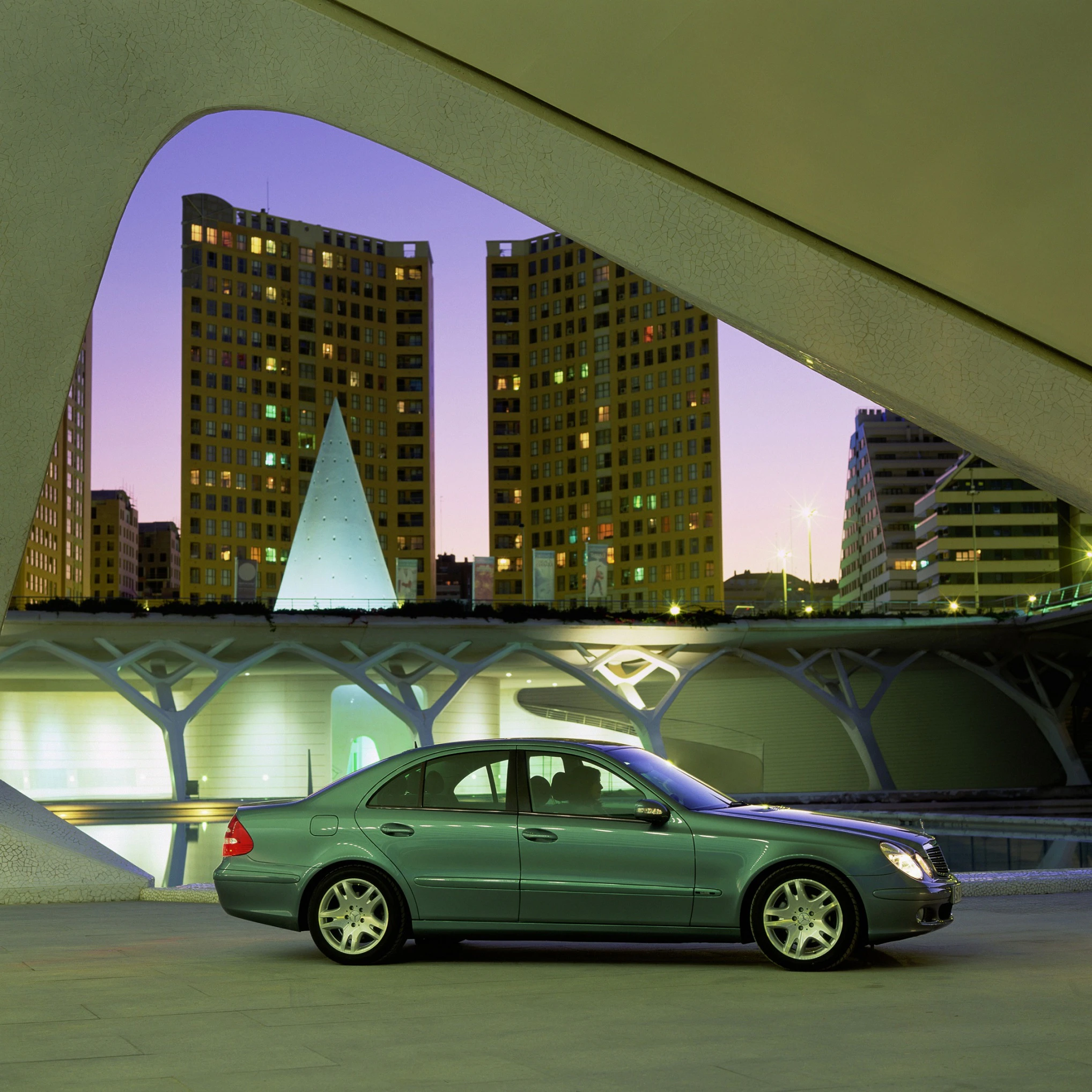 Grünes Auto unter moderner Brücke bei Sonnenuntergang, im Hintergrund beleuchtete Gebäude und ein pyramidenförmiges Bauwerk.