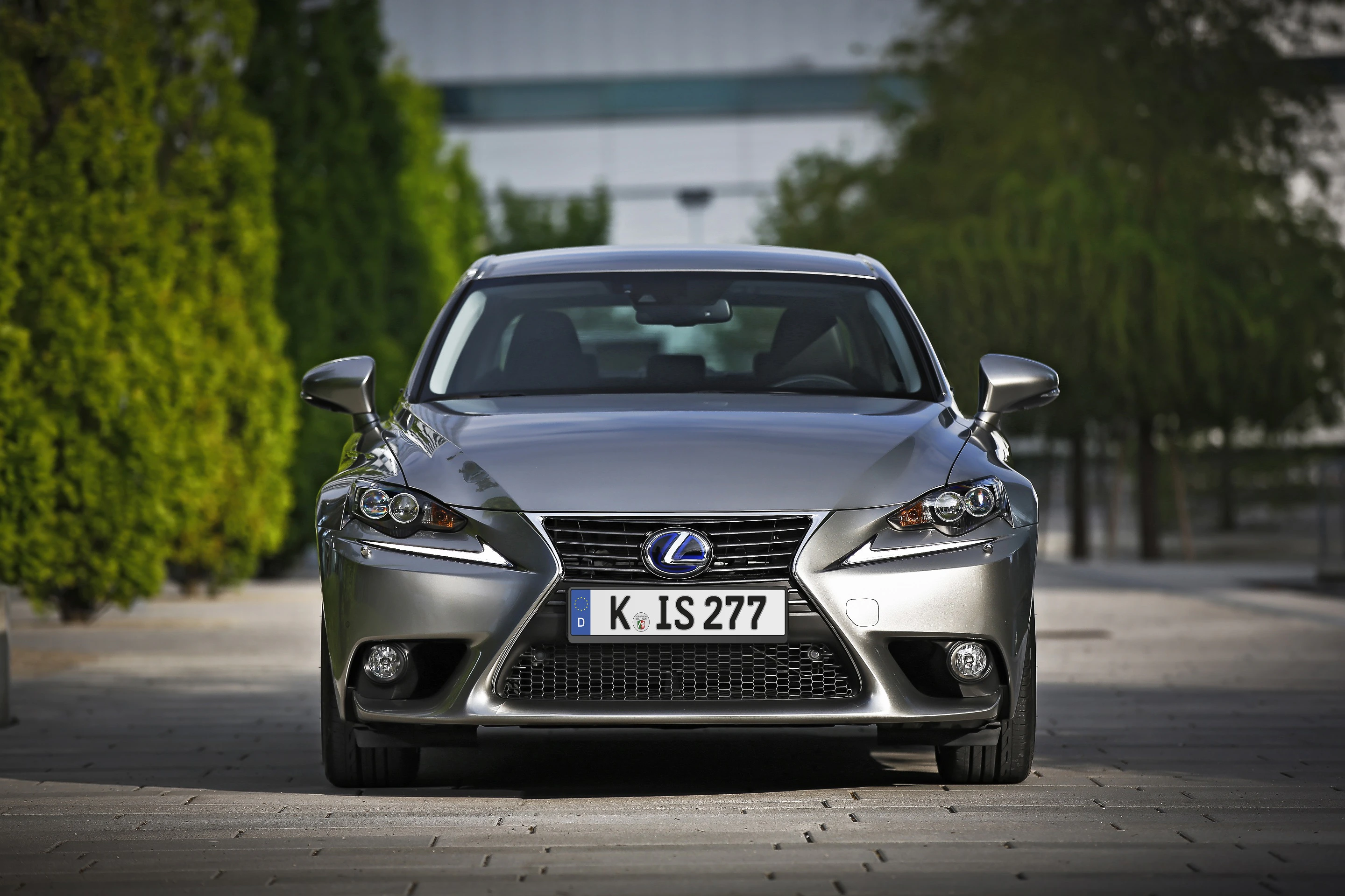 Silbergrauer Lexus IS auf einer gepflasterten Straße, frontal fotografiert, umgeben von grünen Bäumen und einem modernen Gebäude im Hintergrund.