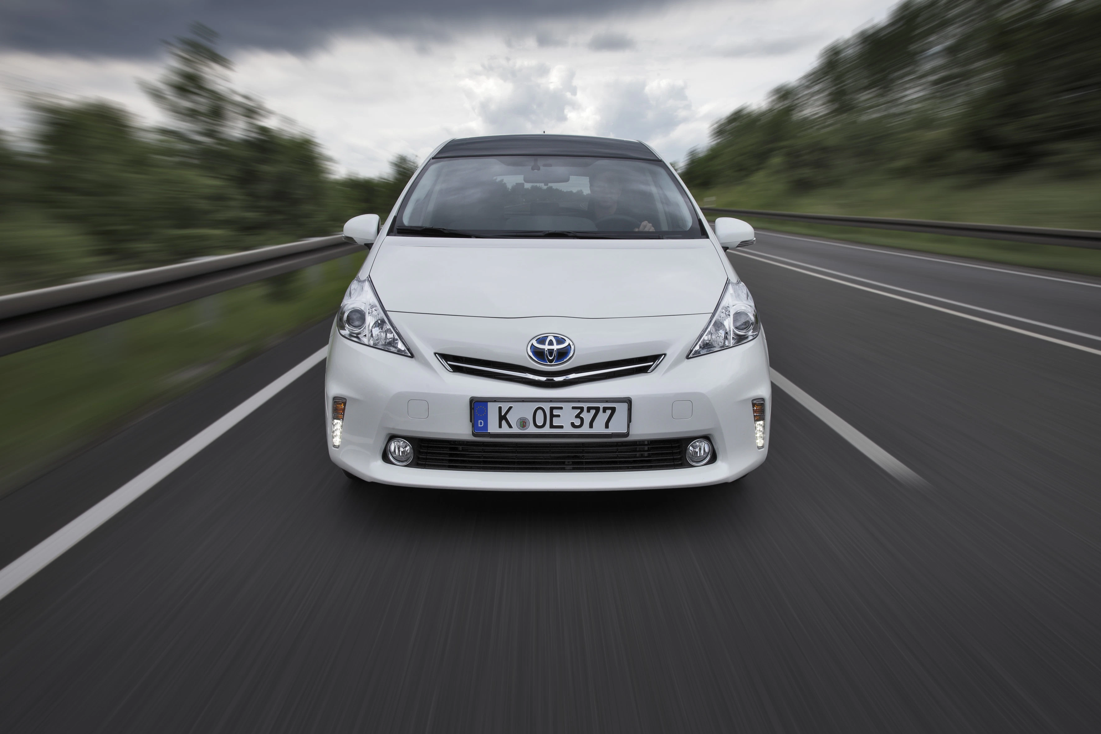 Weißer Toyota Prius fährt auf einer zweispurigen Landstraße, umgeben von verschwommenen Bäumen und einem bewölkten Himmel.