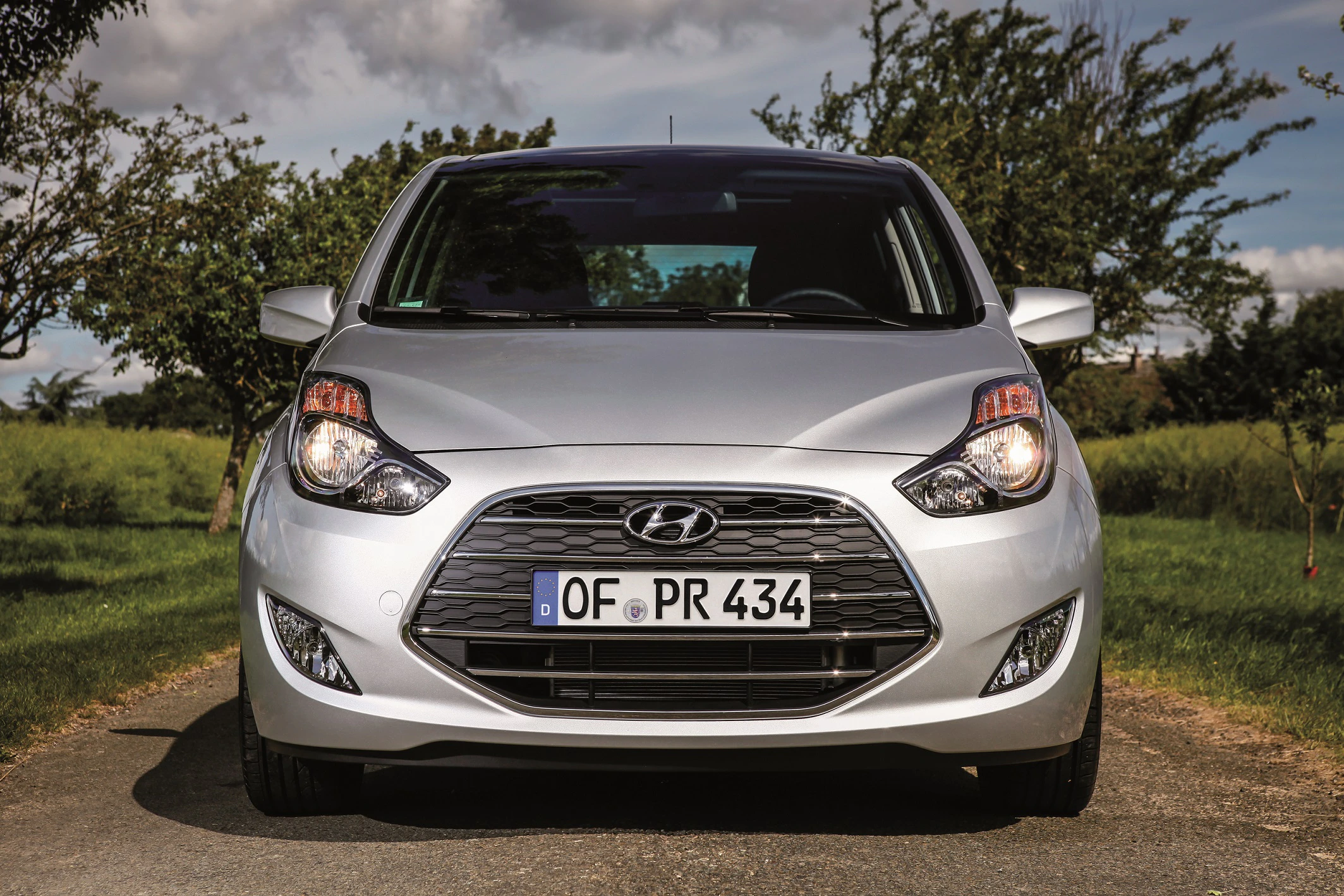 Silberner Hyundai ix20 steht auf einer ländlichen Straße, umgeben von grünen Bäumen und Wiesen, unter klarem Himmel.