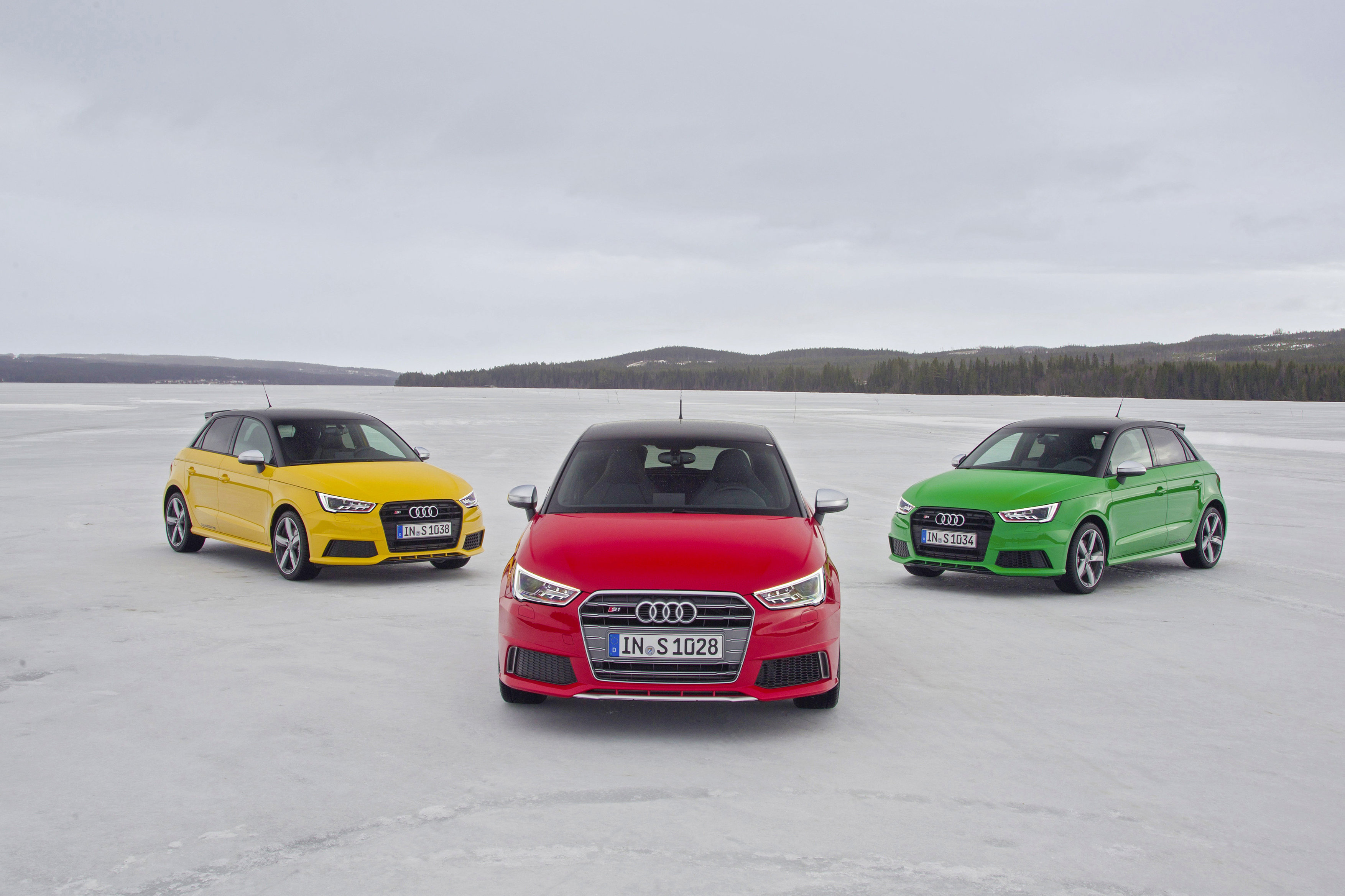 Drei Audi A1 Autos auf schneebedecktem Terrain. Rot in der Mitte, flankiert von gelbem und grünem Auto. Hintergrund: bewaldete Berge und grauer Himmel.
