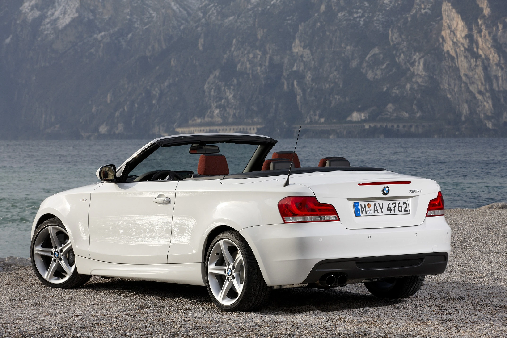 Weißes Cabriolet steht auf einem Kiesstrand vor einer Berg- und Seenlandschaft. Verdeck geöffnet, Nummernschild sichtbar.