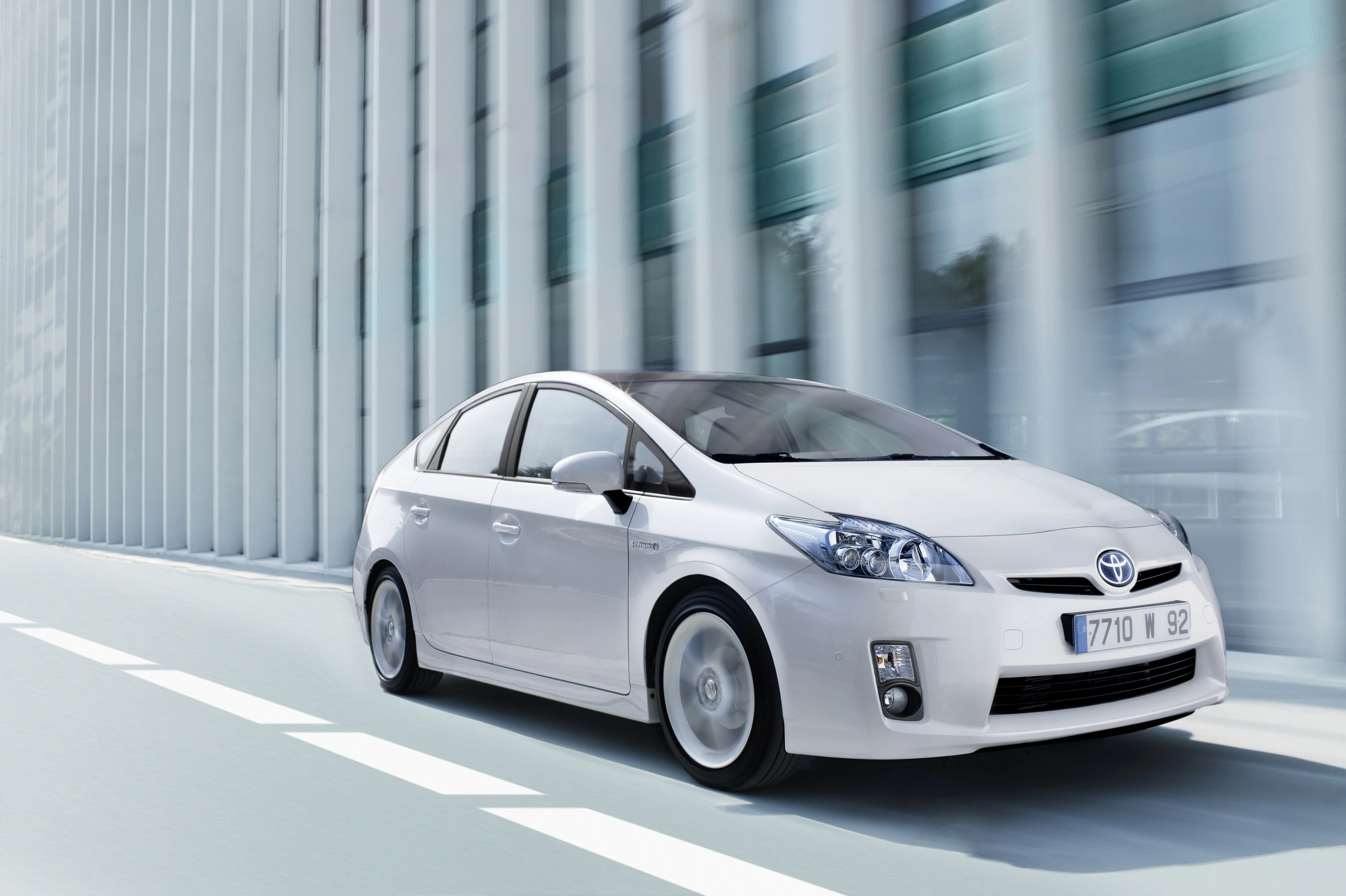 Weißer Toyota Prius fährt dynamisch auf einer Straße mit modernen Gebäuden im Hintergrund.