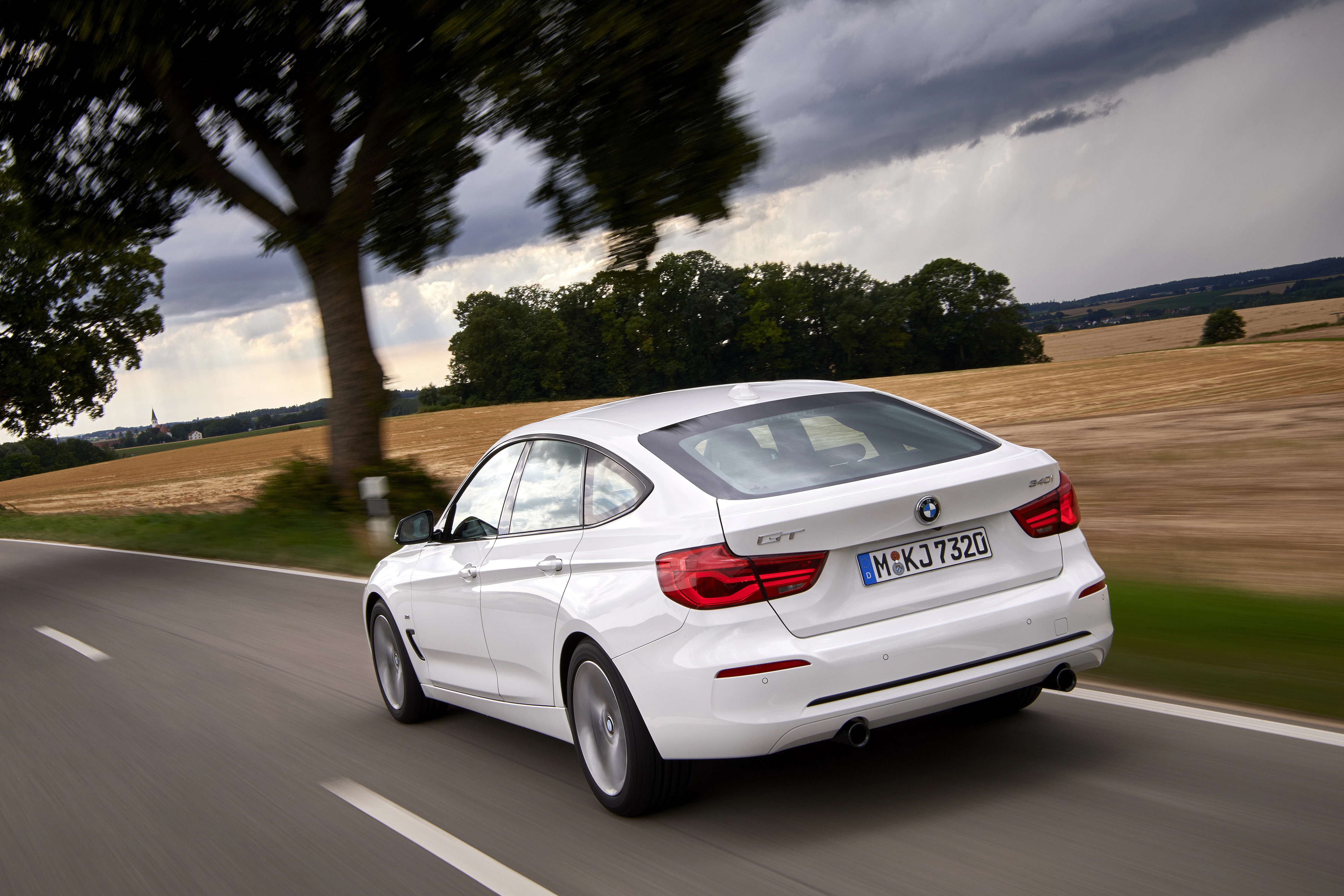 Weißer BMW fährt auf ländlicher Straße, vorbei an Feldern und Bäumen bei bewölktem Himmel.