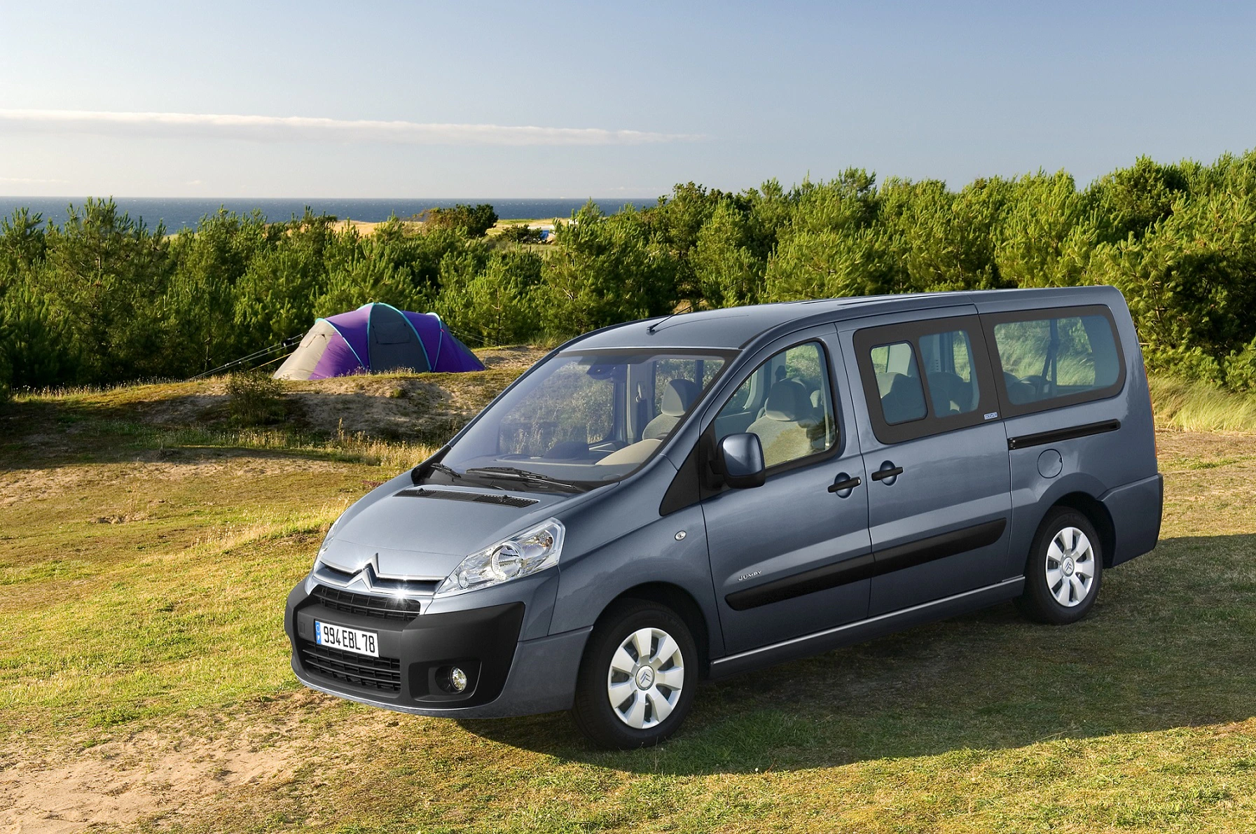 Silberner Citroën-Van parkt auf Wiese, im Hintergrund ein Zelt und Bäume. Meer am Horizont unter klarem Himmel.