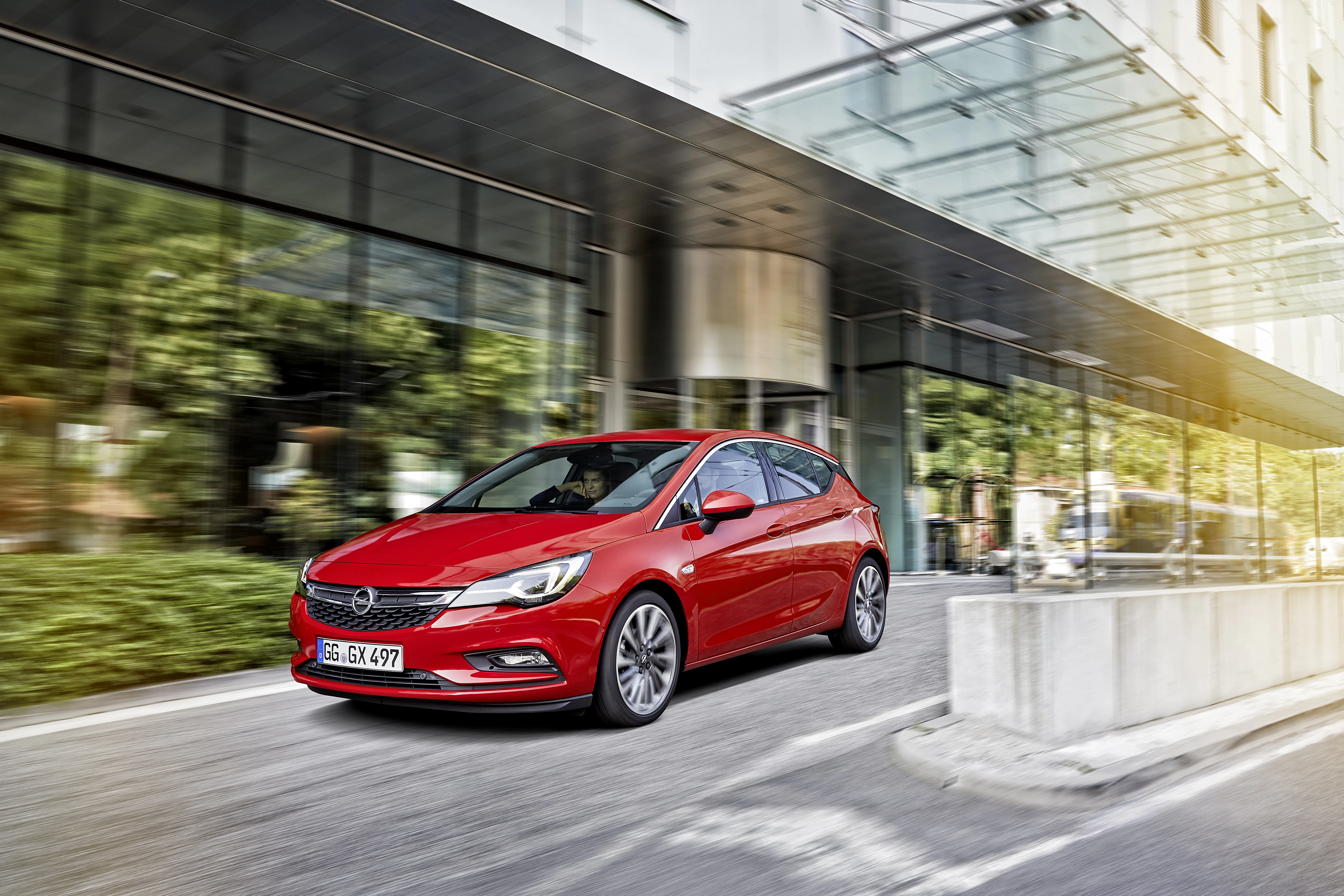 Roter Opel Astra fährt unter Glasdach vor modernem Gebäude, Bäume spiegeln sich in den Fenstern.