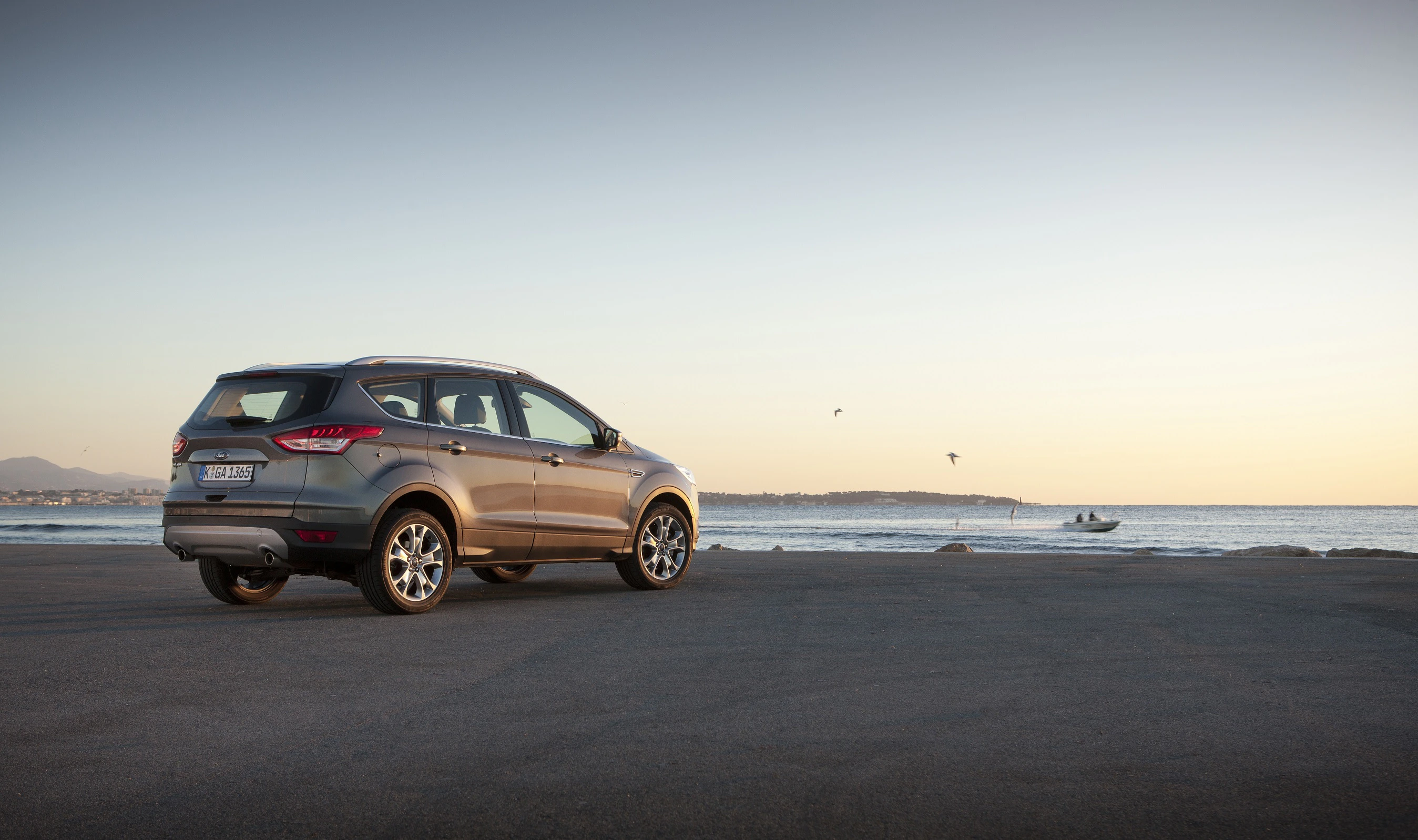 Silberner SUV auf leerem Parkplatz mit Blick auf das Meer bei Sonnenuntergang. Vögel fliegen, kleines Boot am Horizont sichtbar.