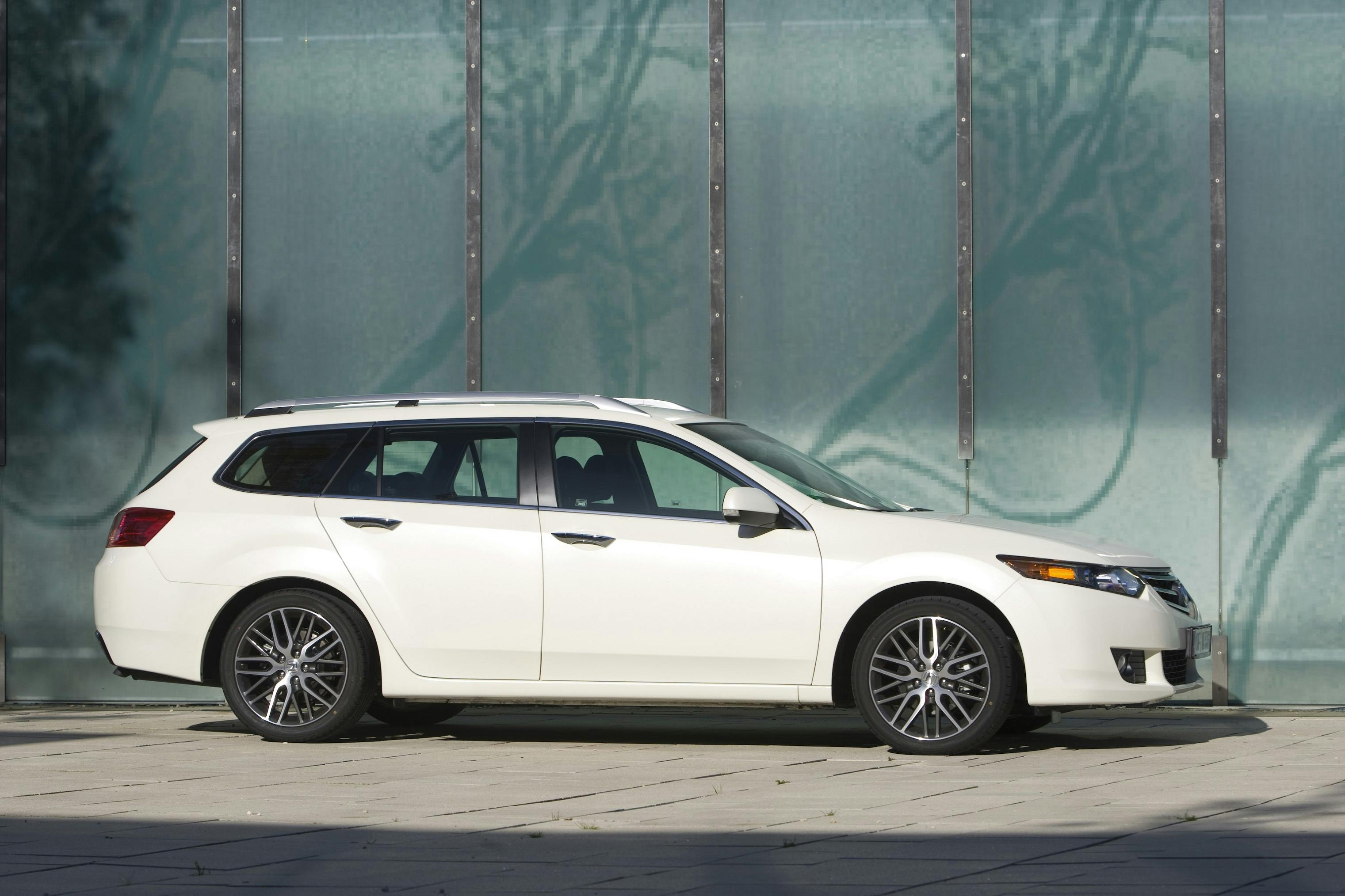 Weißer Kombi parkt vor einer modernen Glaswand. Das Auto hat polierte Felgen und ist seitlich fotografiert.