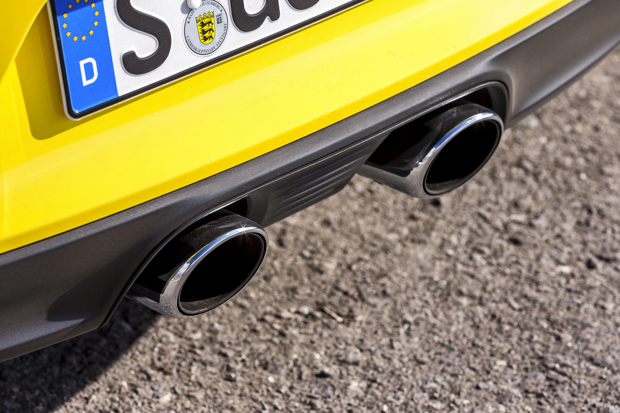Nahaufnahme eines gelben Autos mit zwei verchromten Auspuffrohren und einem Teil des Nummernschilds auf einer asphaltierten Straße.