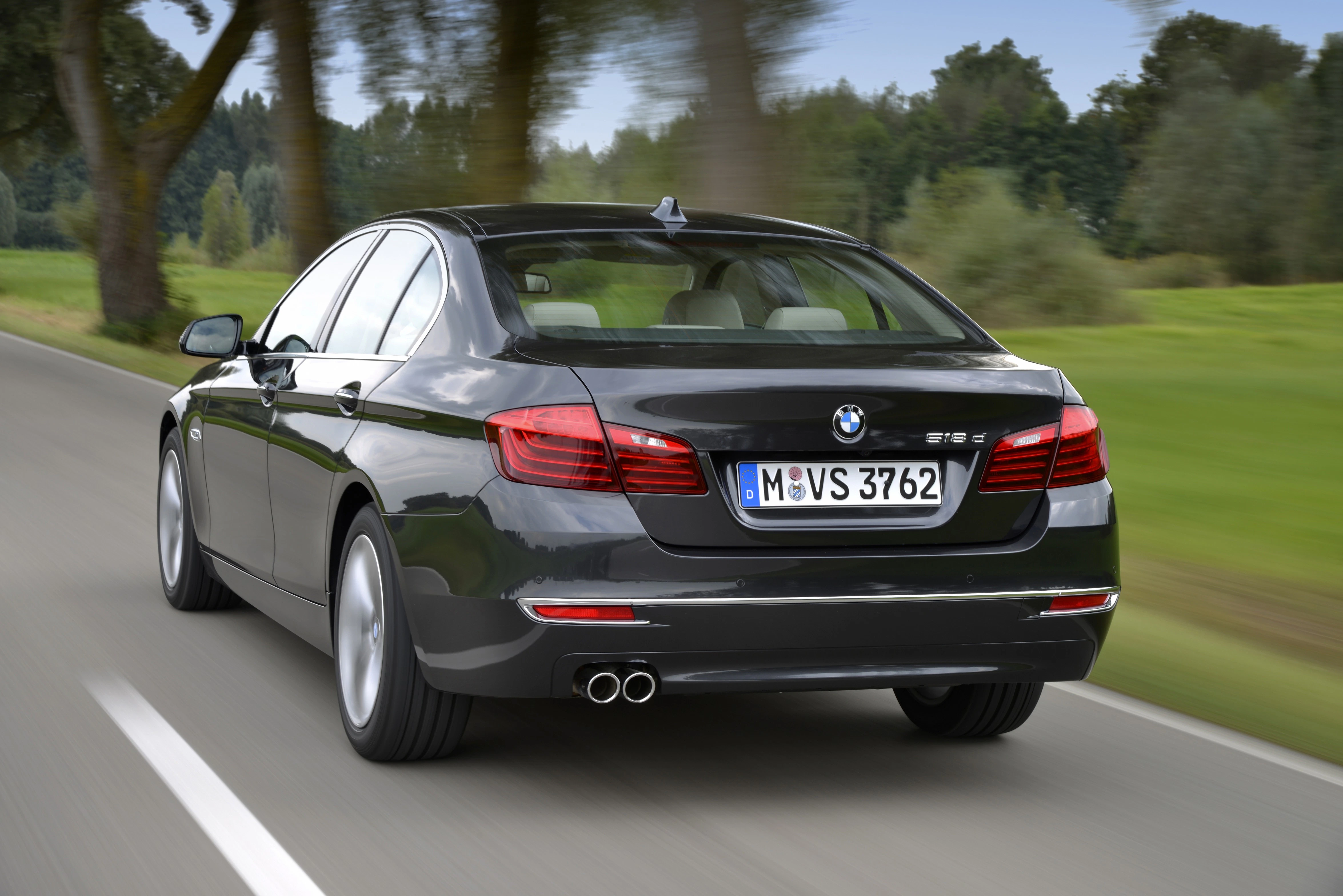 Dunkelgrauer BMW 5er Limousine fährt auf einer Landstraße mit Bäumen und Wiesen im Hintergrund. Straßenschild mit Biegung voraus.