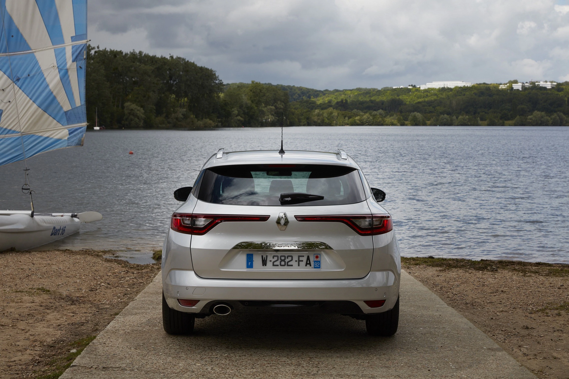 Silbernes Auto steht rückwärts am Seeufer, neben einem Segelboot. Bewölkter Himmel und bewaldete Landschaft im Hintergrund.