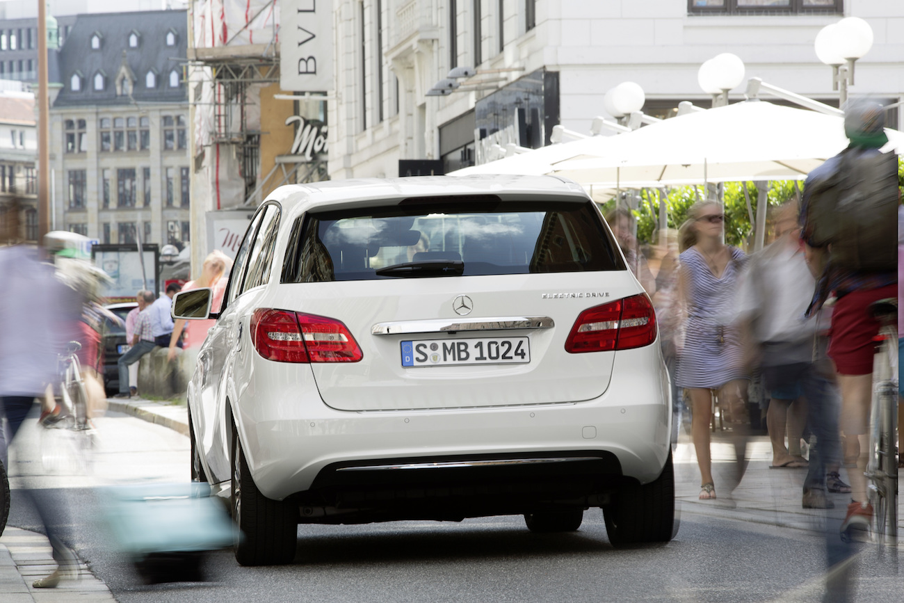 Weißer Mercedes-Benz fährt durch belebte, städtische Straße. Passanten und Radfahrer bewegen sich dynamisch um das Auto herum.