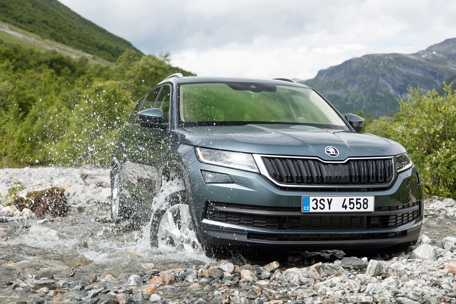 Ein grauer SUV fährt durch einen flachen Bach in einer bergigen Landschaft. Spritzendes Wasser umgibt das Fahrzeug.