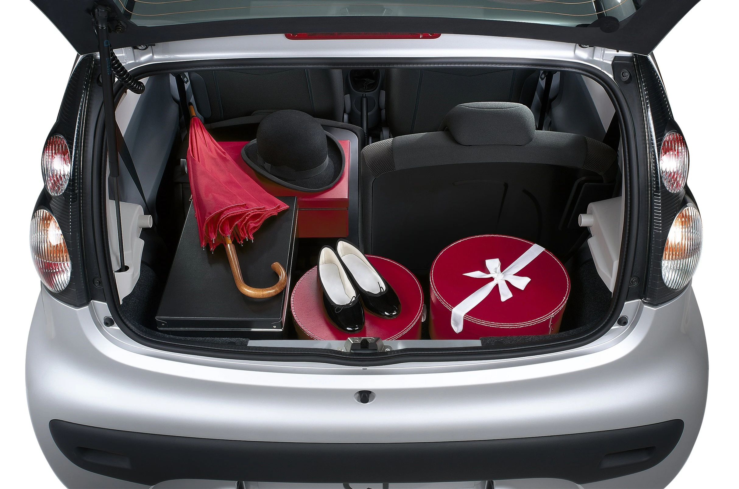Kofferraum eines Autos mit einem roten Regenschirm, schwarzem Hut, weißen Schuhen und runden, roten Hutboxen.