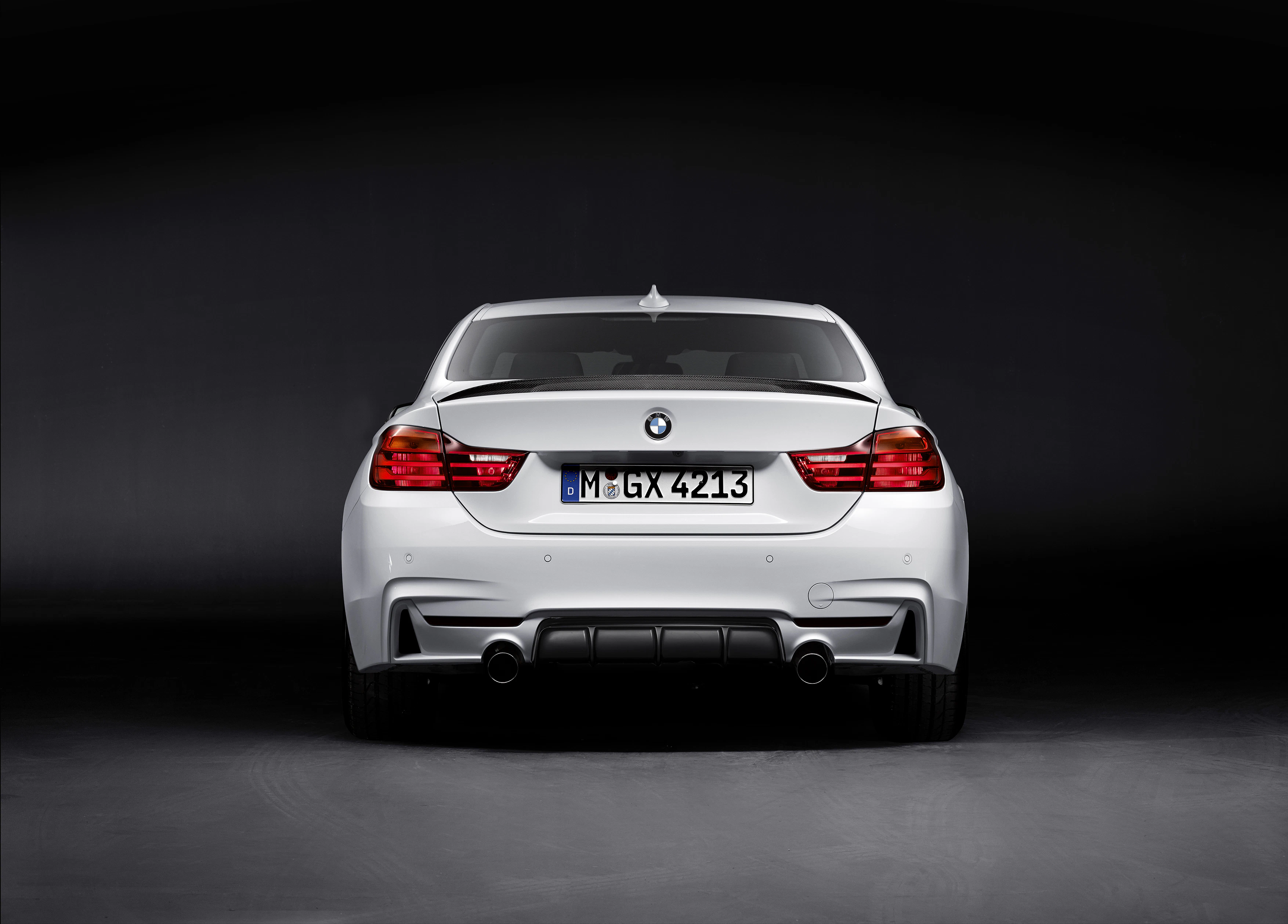 Rückansicht eines weißen BMW M4 mit Kennzeichen, stehend auf dunklem Hintergrund. Modernes Design mit klaren Linien und Doppelauspuff.