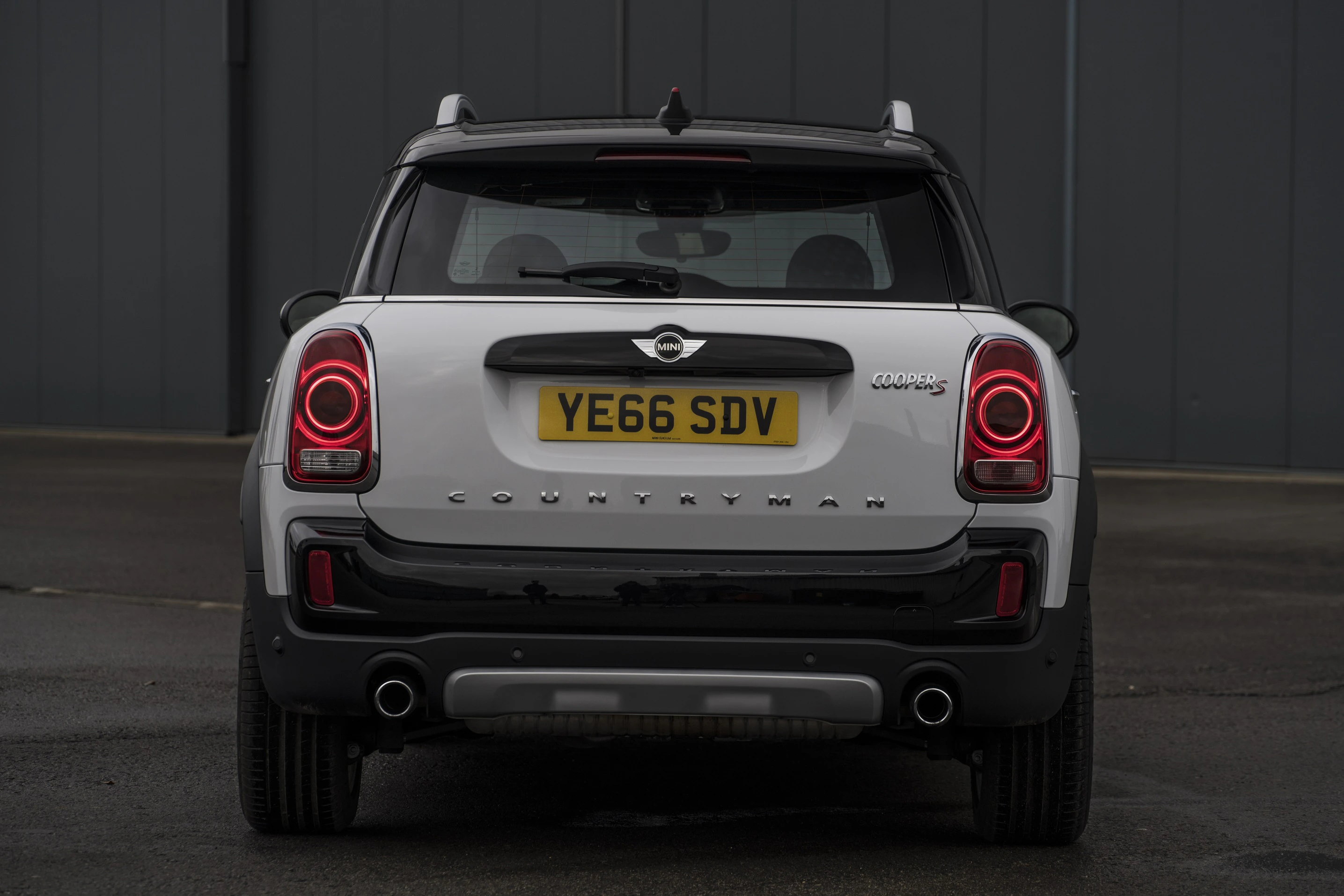 Rückansicht eines weißen Mini Countryman mit schwarzem Dach, rotem Rücklicht und britischem Kennzeichen auf dunklem Hintergrund.