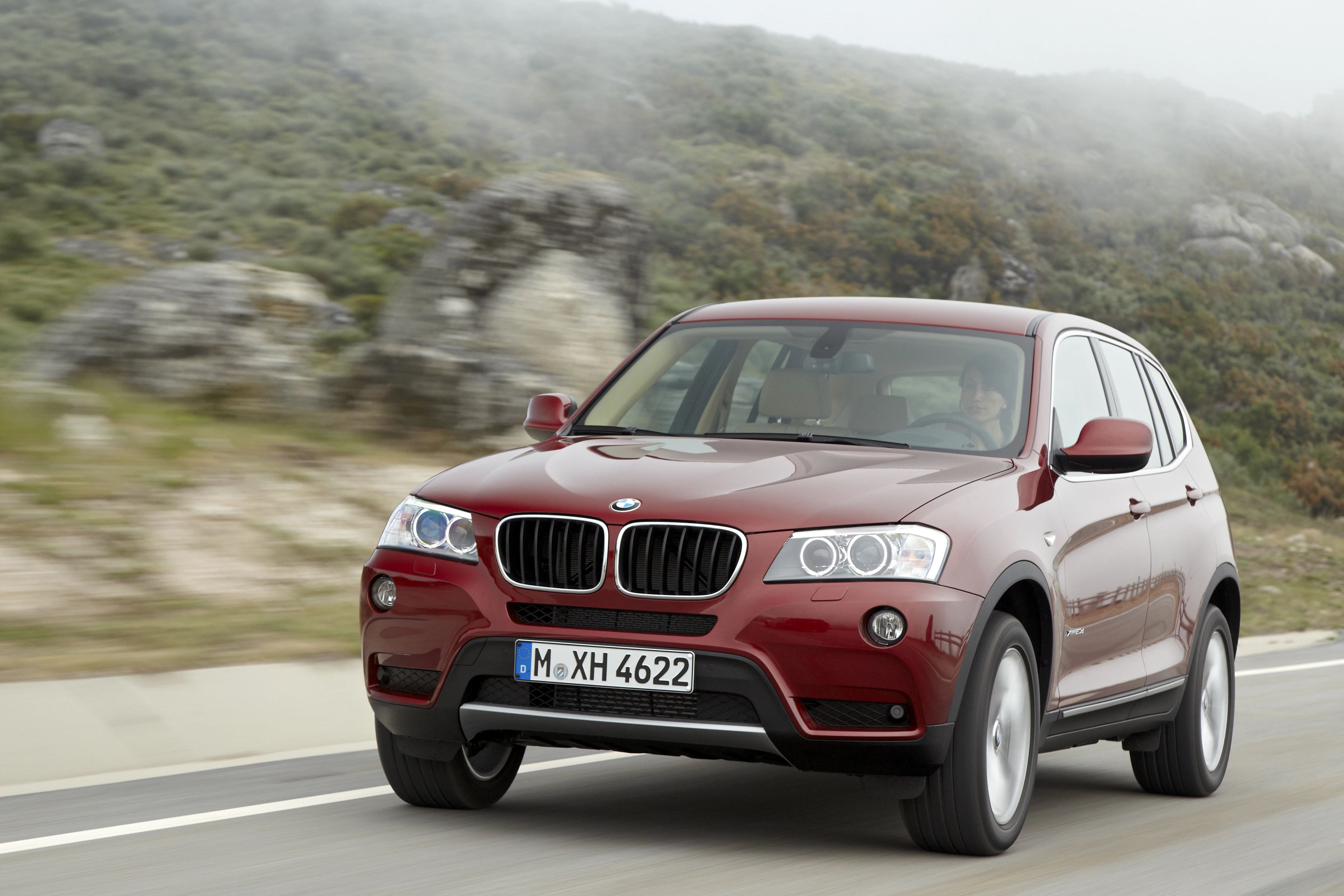 Roter BMW SUV fährt auf einer Landstraße, im Hintergrund sind bewaldete Hügel und ein bewölkter Himmel zu sehen.