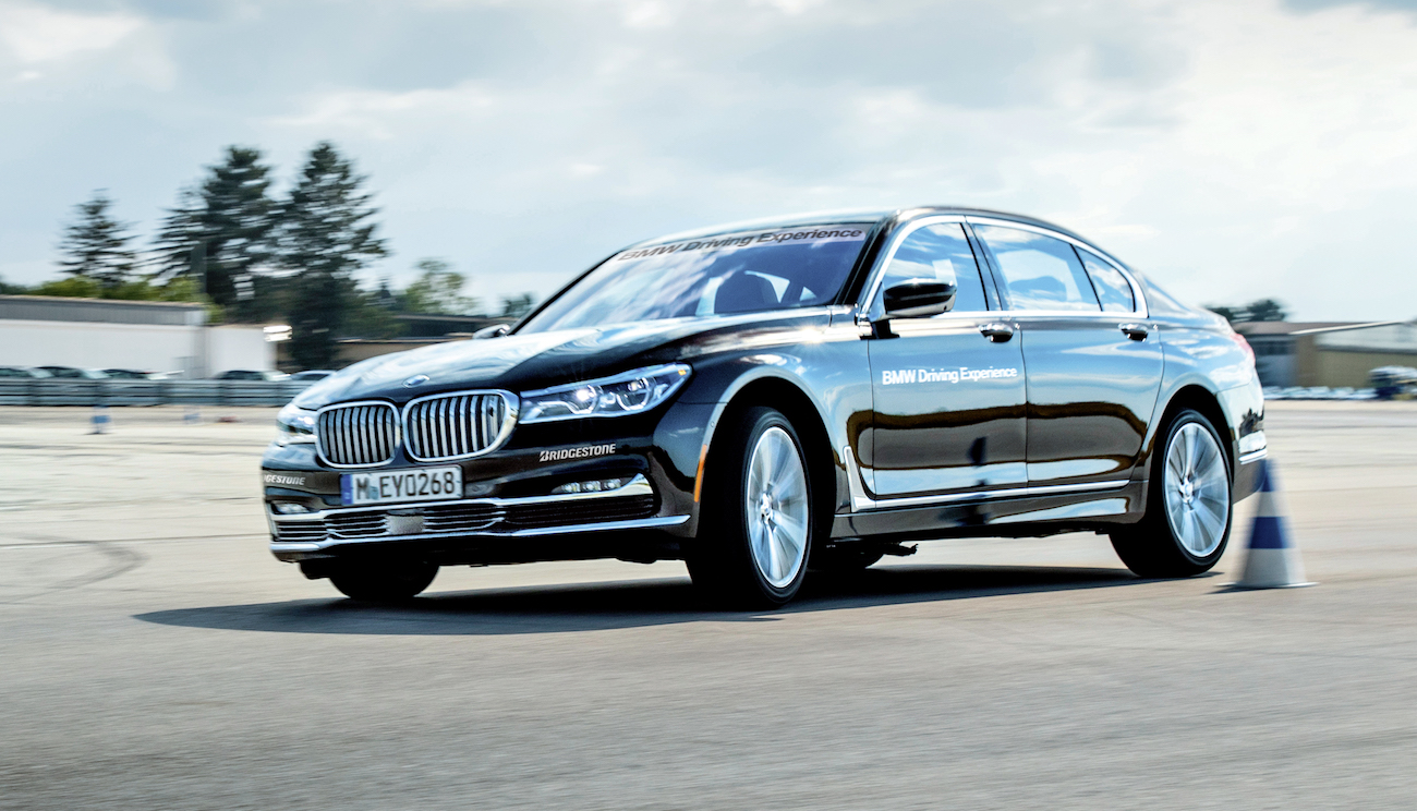 BMW-Limousine fährt dynamisch auf einer Teststrecke mit blauen Hütchen, Gebäude und Bäume im Hintergrund.