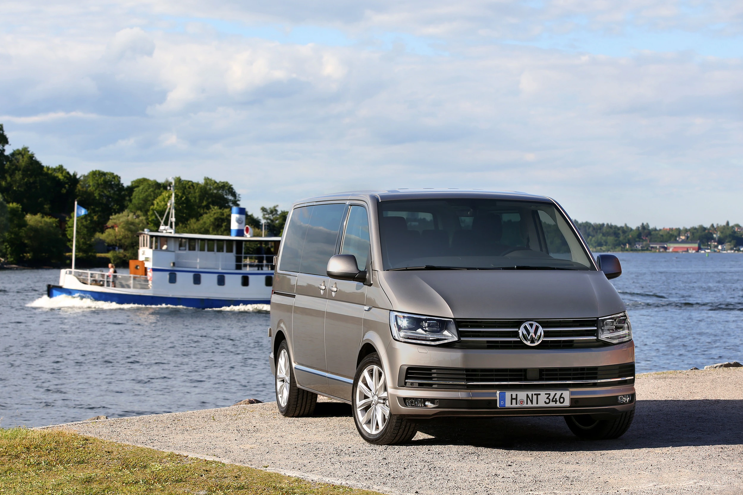 Silberner VW Transporter am Ufer eines Sees, im Hintergrund fährt ein blau-weißes Schiff. Klarer Himmel mit vereinzelten Wolken.
