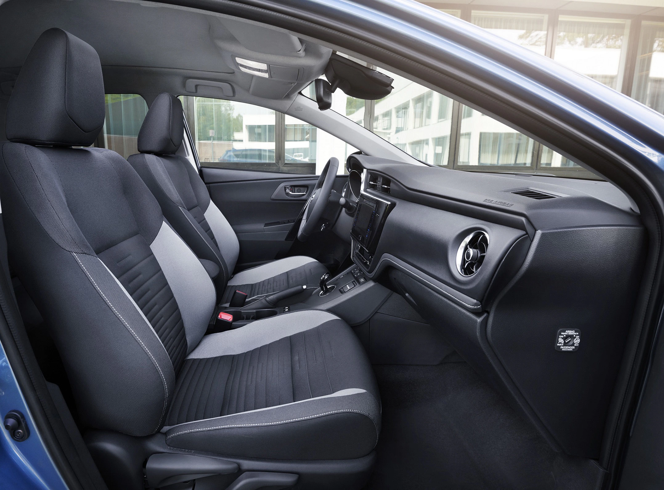 Innenraum eines modernen Autos mit zwei Vordersitzen, grauen Stoffbezügen und einem Armaturenbrett mit integriertem Bildschirm.
