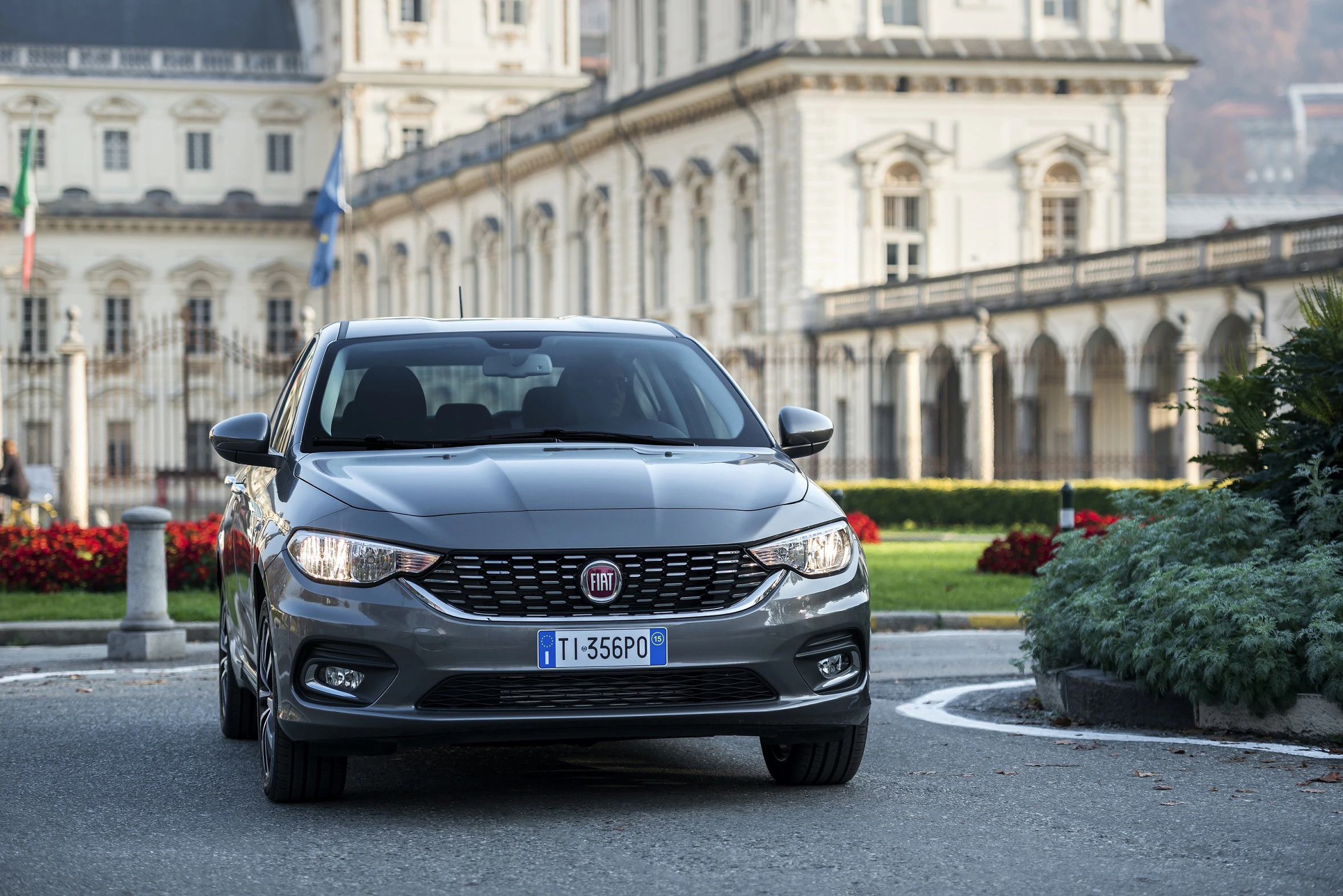 Graues Auto (Fiat) vor historischem Gebäude mit klassischer Architektur, umgeben von bunten Blumen und Bäumen.