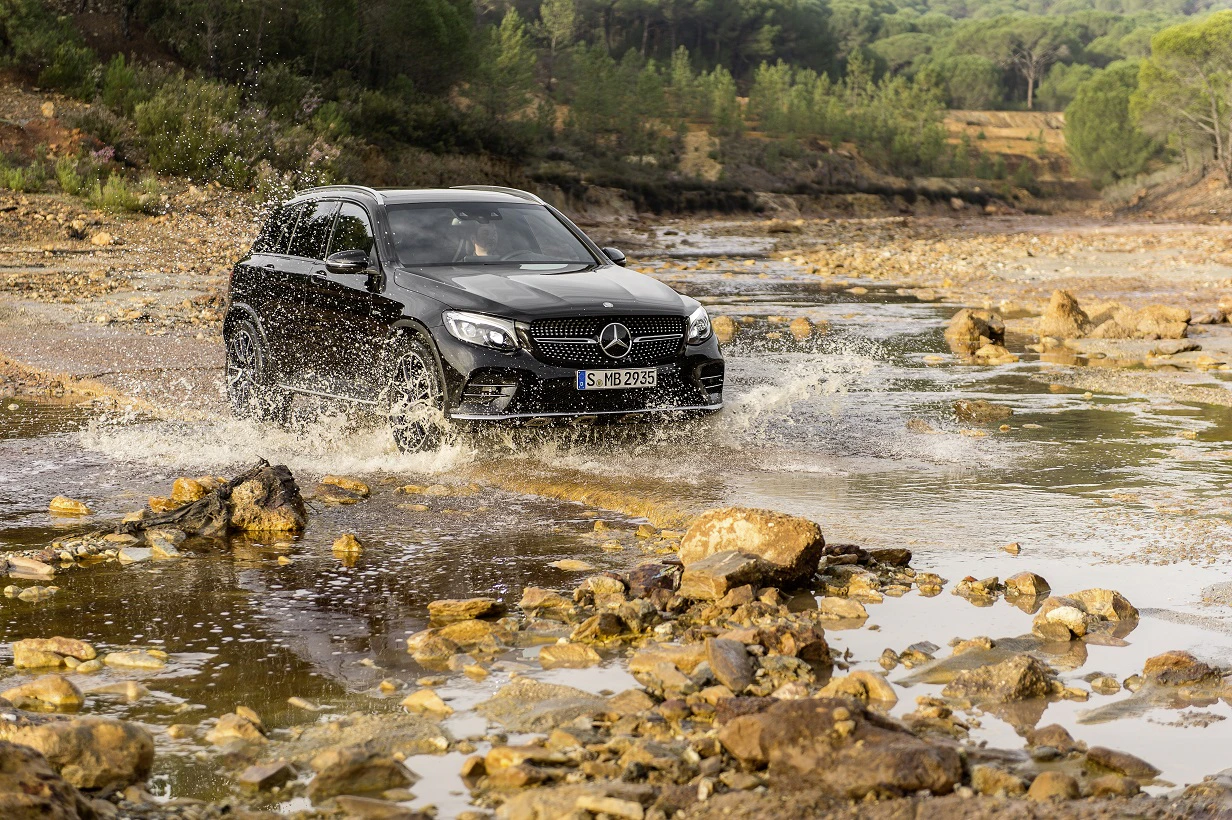 Ein schwarzer SUV fährt durch ein flaches, steiniges Wasserbett in einer natürlichen Landschaft mit grünen Bäumen im Hintergrund.