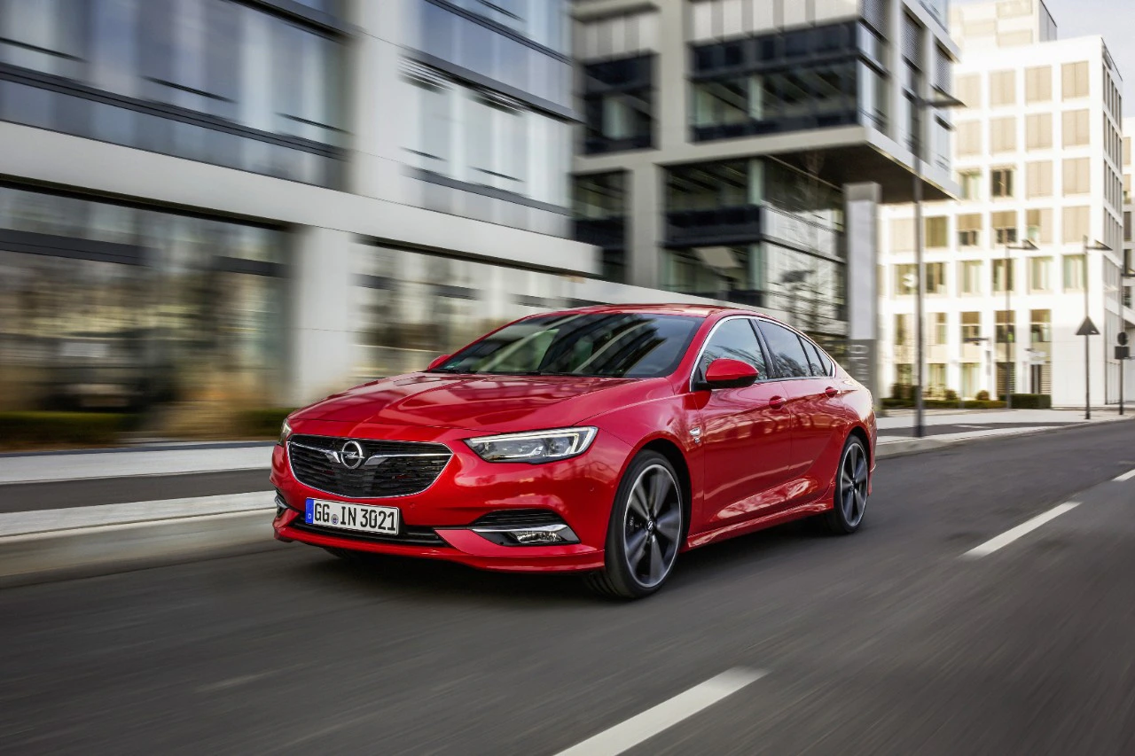 Roter Opel Insignia fährt dynamisch auf einer städtischen Straße, moderne Gebäude im Hintergrund.