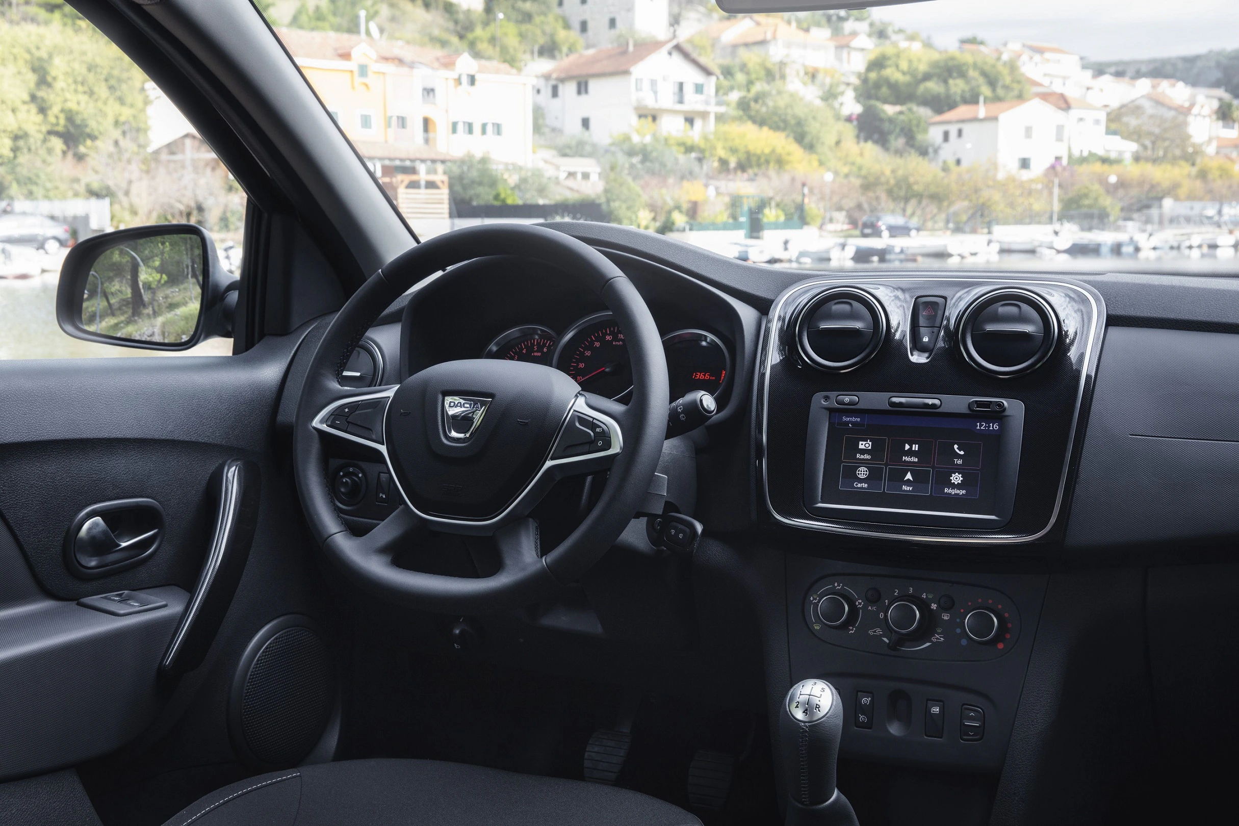 Innenraum eines Autos mit Lenkrad, Armaturenbrett und Touchscreen-Display. Gebäude und Natur im Hintergrund durch das Fenster sichtbar.