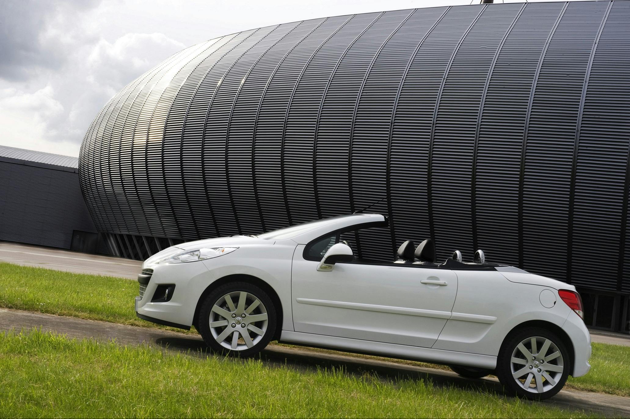 Weißes Cabrio-Peugeot parkt vor einem modernen, gebogenen Gebäude aus Metall mit Wolken im Hintergrund.