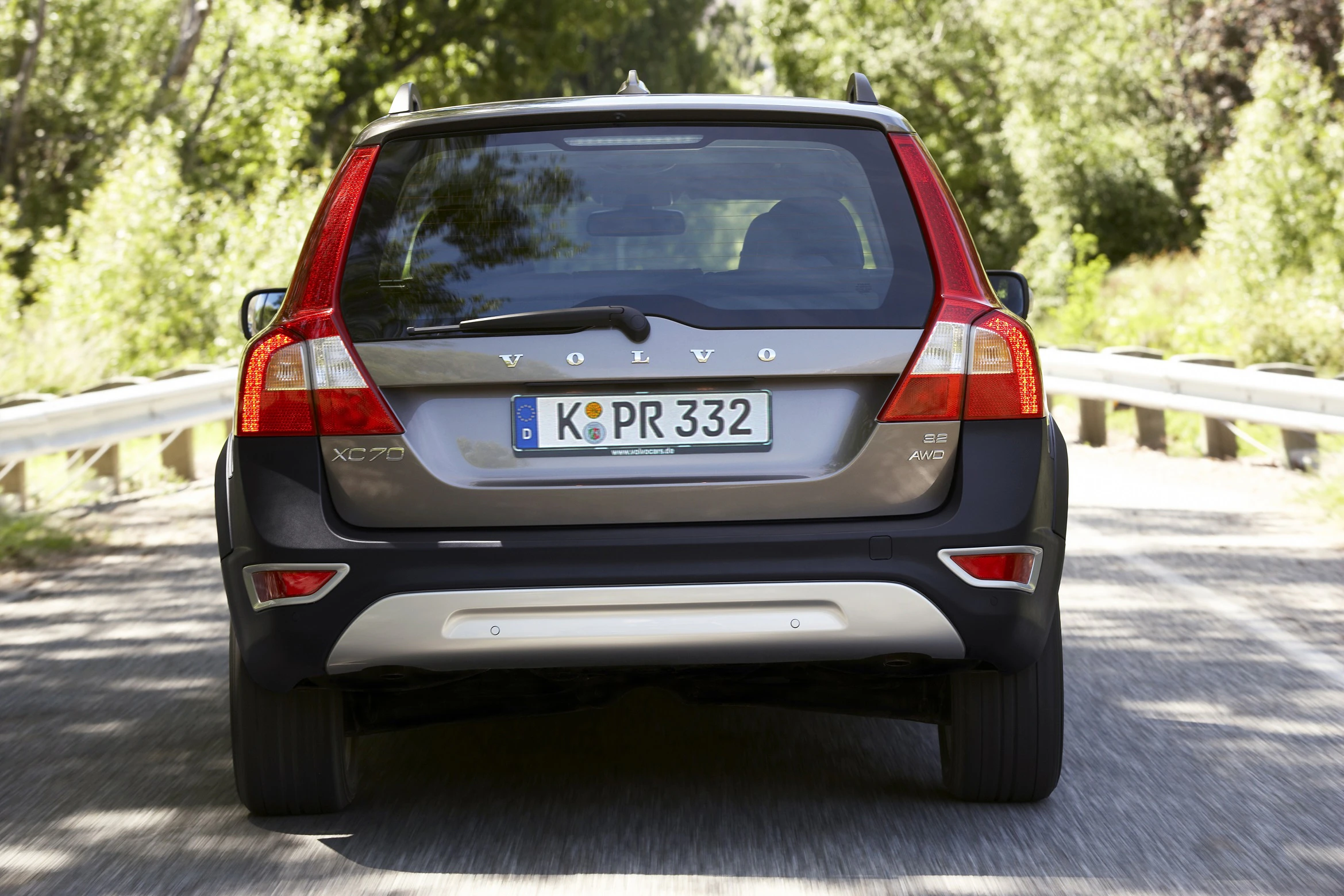 Ein grauer Volvo XC70 fährt auf einer sonnigen Landstraße, umgeben von grüner Vegetation. Das Auto hat ein deutsches Nummernschild.
