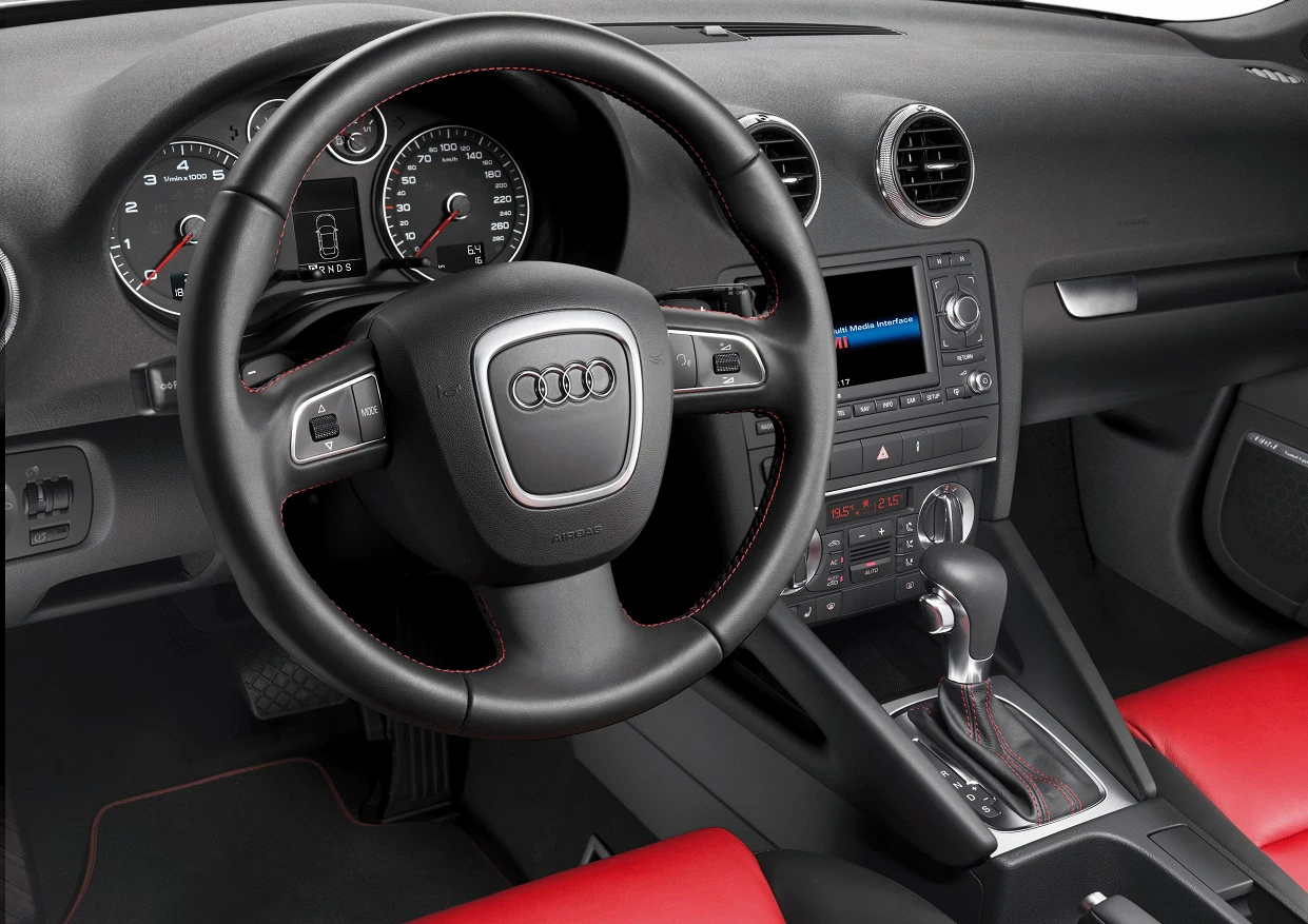 Nahaufnahme eines Audi-Innenraums mit schwarzem Lenkrad, roter Innenausstattung, Tachometer und zentralem Display. Automatikgetriebe erkennbar.