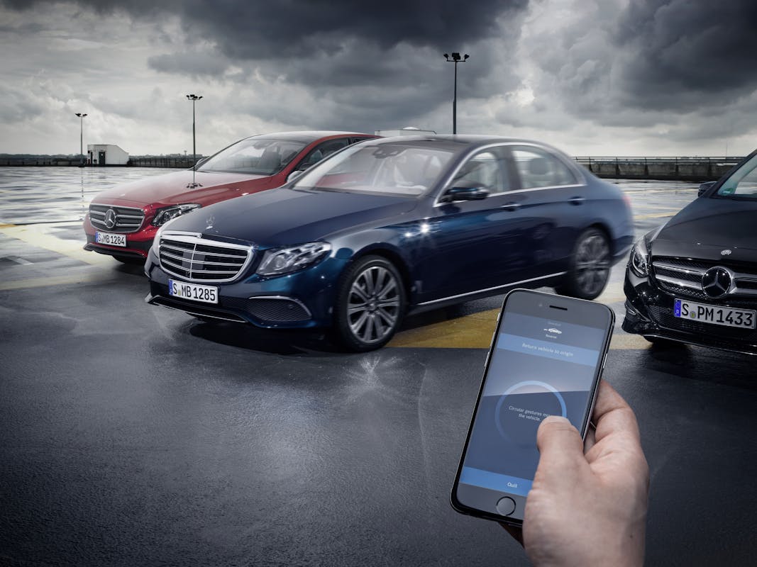 Person hält Smartphone vor drei parkenden Mercedes-Benz Autos auf nasser Fläche mit bewölktem Himmel im Hintergrund.