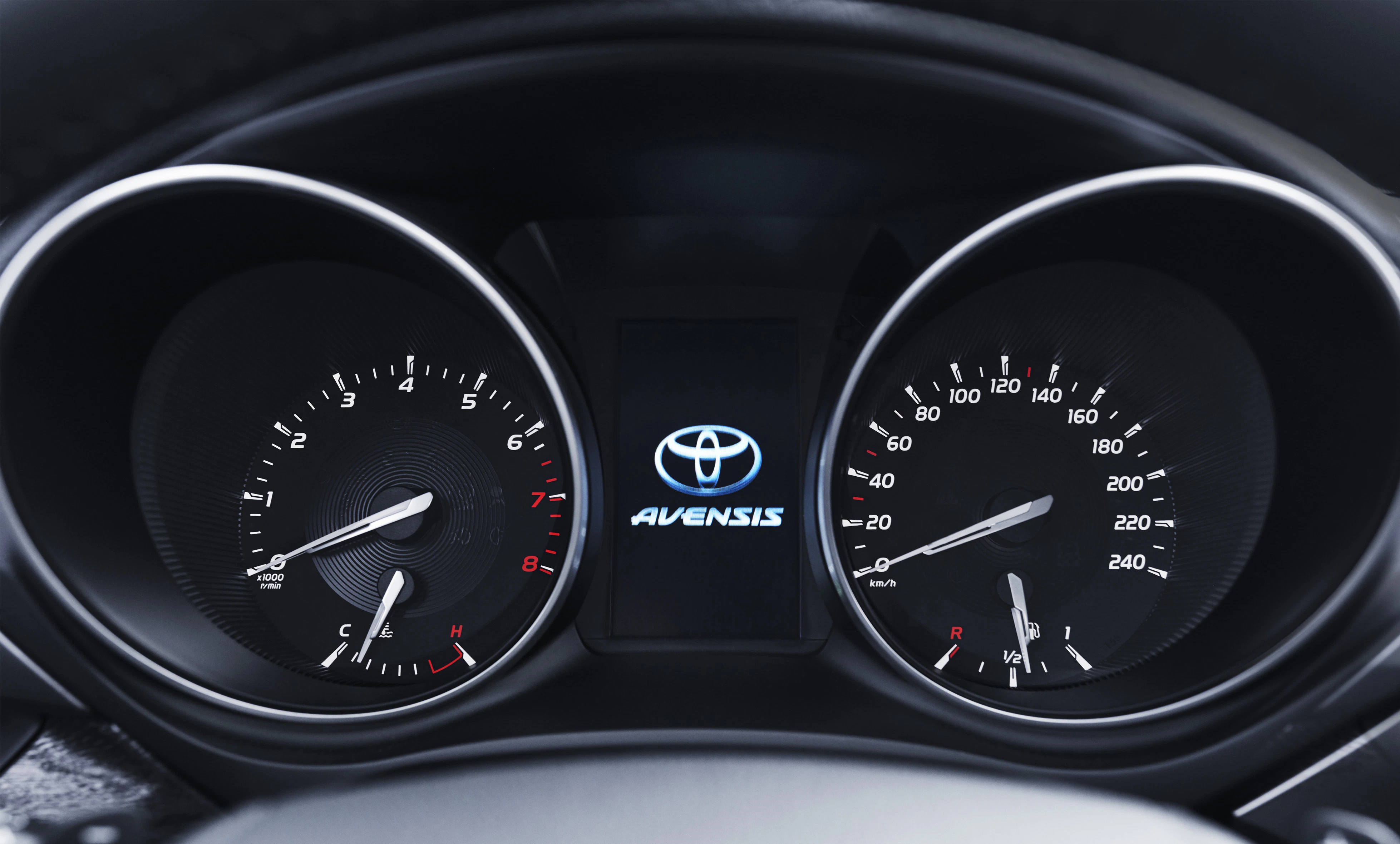 Nahaufnahme eines Toyota Avensis Armaturenbretts mit Tachometer, Drehzahlmesser und zentralem Display mit Toyota-Logo.