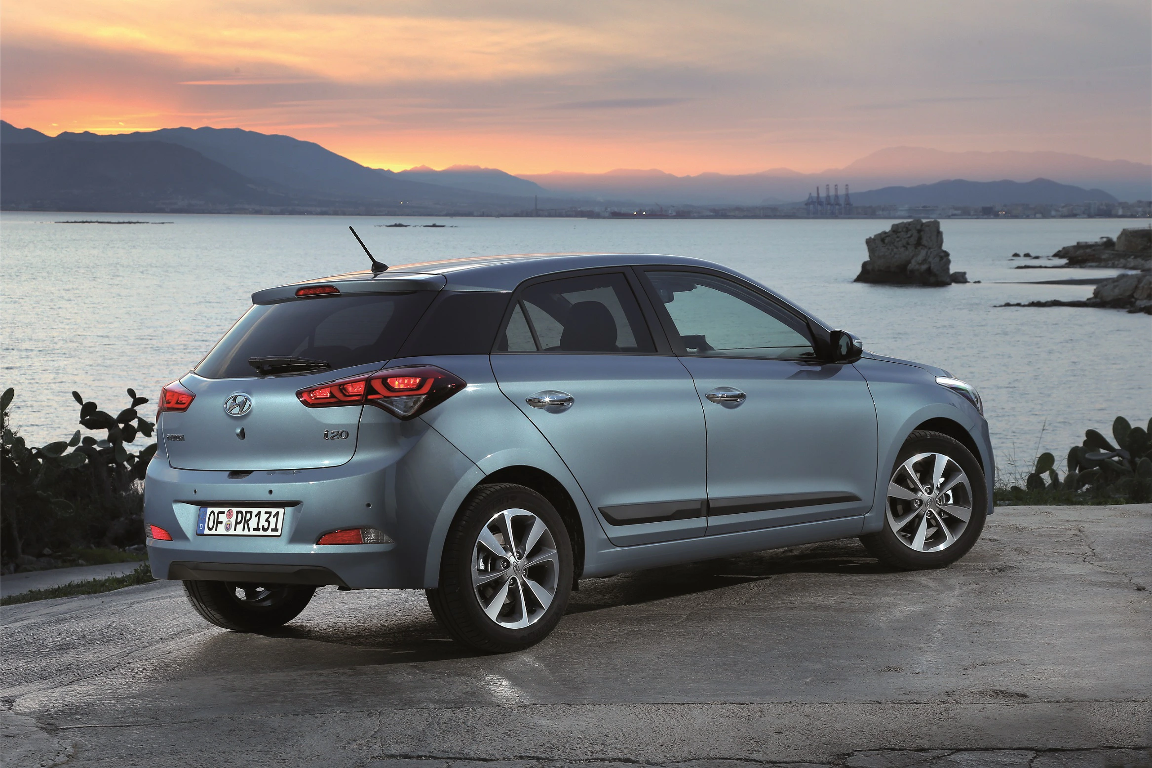 Silberner Hyundai i20 steht am Meer bei Sonnenuntergang. Im Hintergrund sind Berge und das ruhige Wasser des Meeres zu sehen.