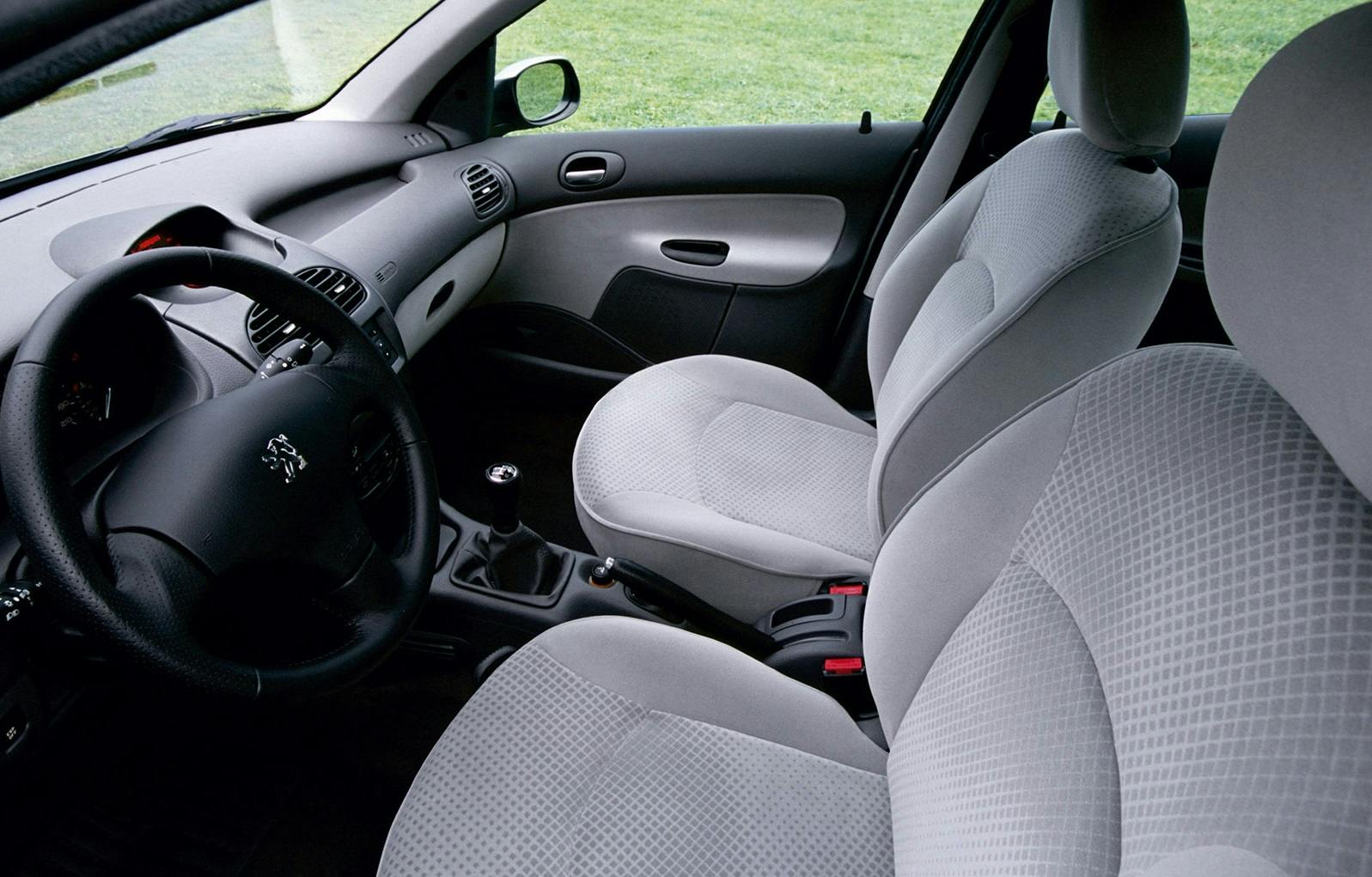 Innenraum eines Autos mit grauen Stoffpolstern, manueller Gangschaltung und schwarzem Lenkrad. An einer Wiese geparkt.