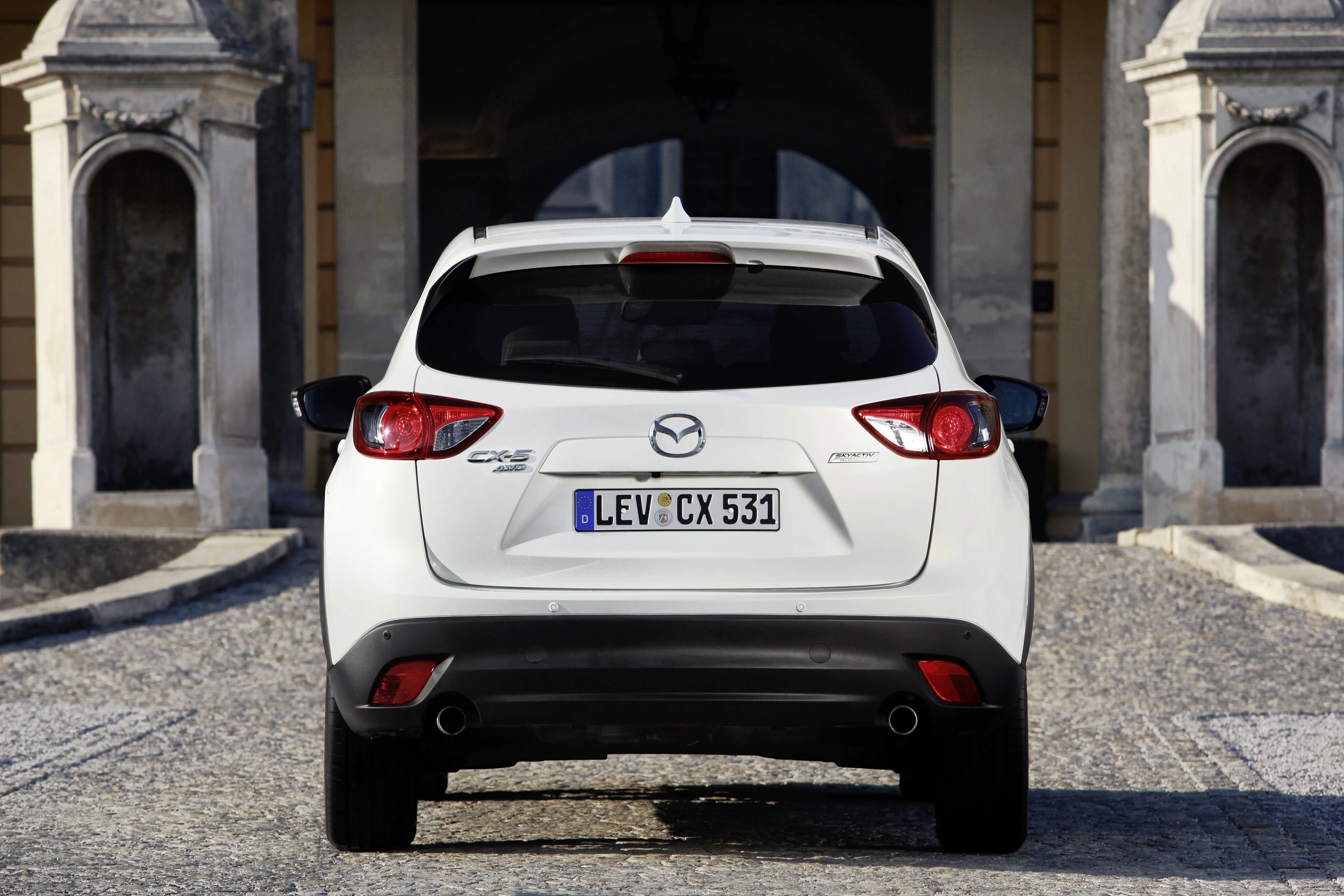 Rückansicht eines weißen Mazda CX-5 mit deutschem Kennzeichen, geparkt vor einem Gebäude mit steinernem Torbogen.