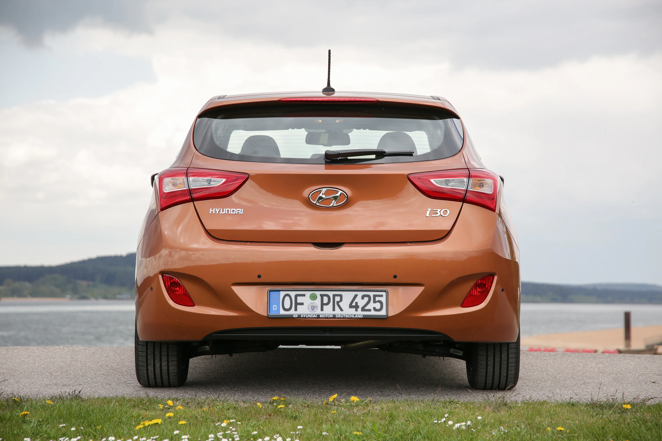 Rückansicht eines orangefarbenen Hyundai i30, geparkt vor einer natürlichen Kulisse mit See und bewölktem Himmel.