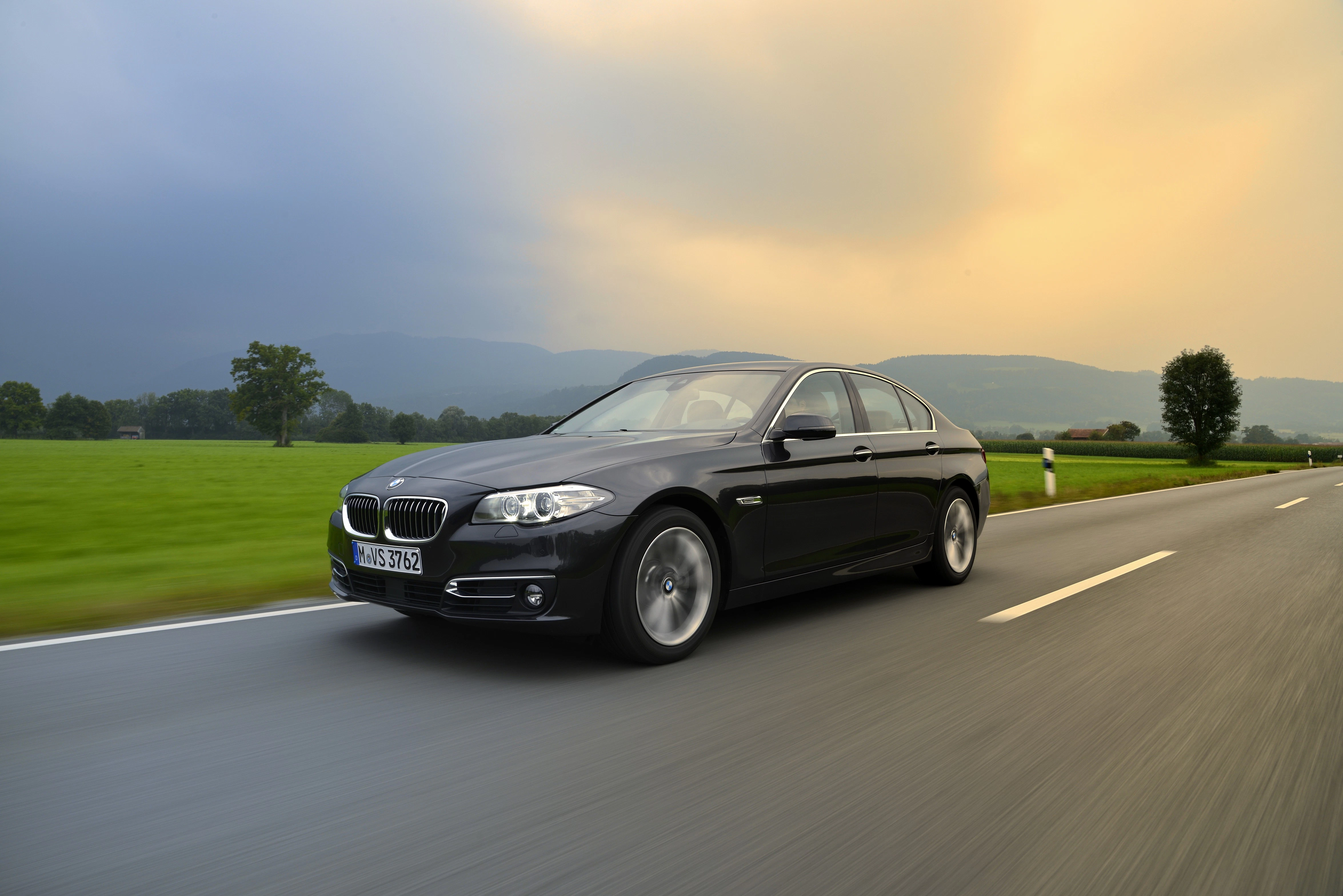 Schwarzer BMW fährt auf einer Landstraße bei Sonnenuntergang, umgeben von grünen Feldern und einem bewölkten Himmel im Hintergrund.