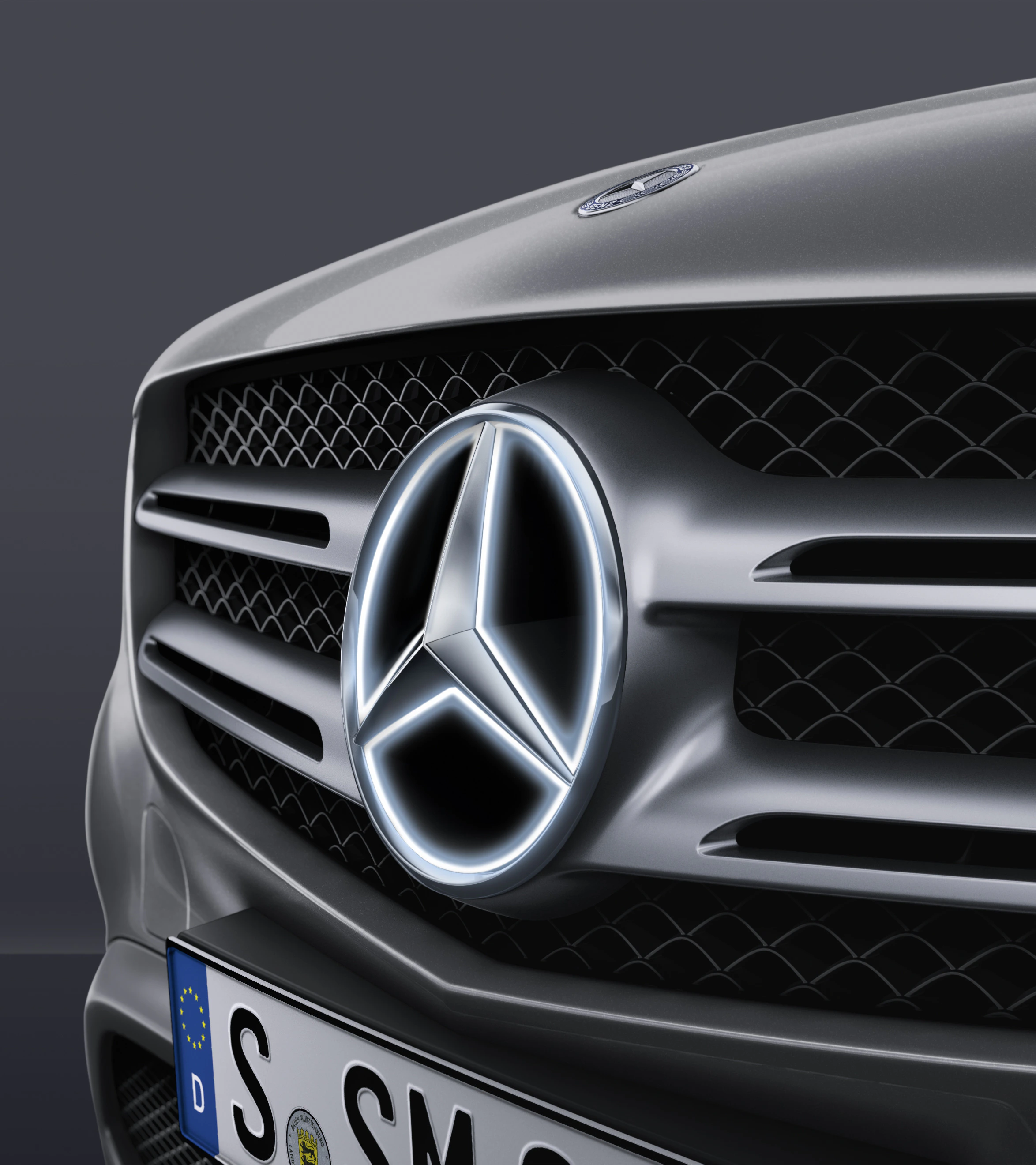 Nahaufnahme eines silbernen Mercedes-Autos mit beleuchtetem Mercedes-Stern auf dem Kühlergrill und deutschem Kennzeichen.