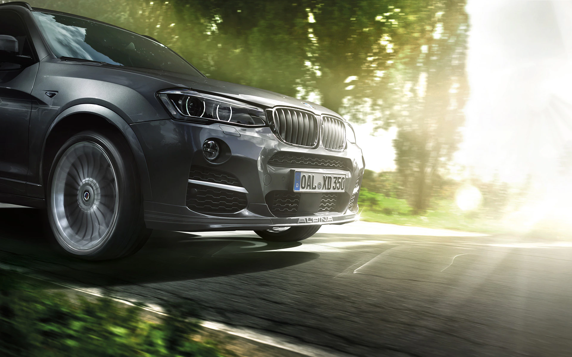 Nahaufnahme eines dunkelgrauen BMW SUV auf einer Landstraße, umgeben von grünen Bäumen und Sonnenstrahlen im Hintergrund.