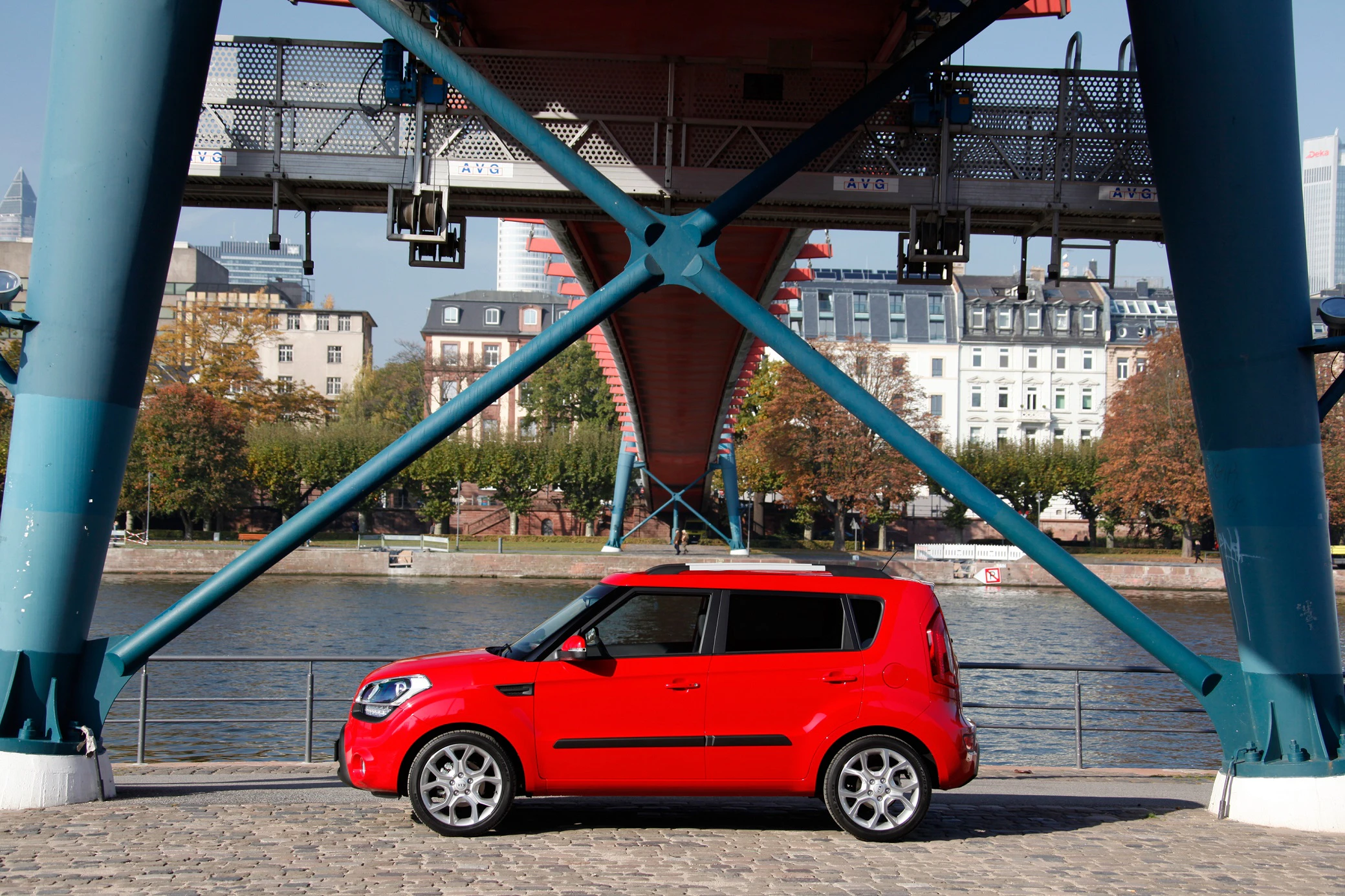 Rotes Auto parkt unter einer stählernen Brücke an einem Fluss, im Hintergrund Bäume und Gebäude bei klarem Himmel.