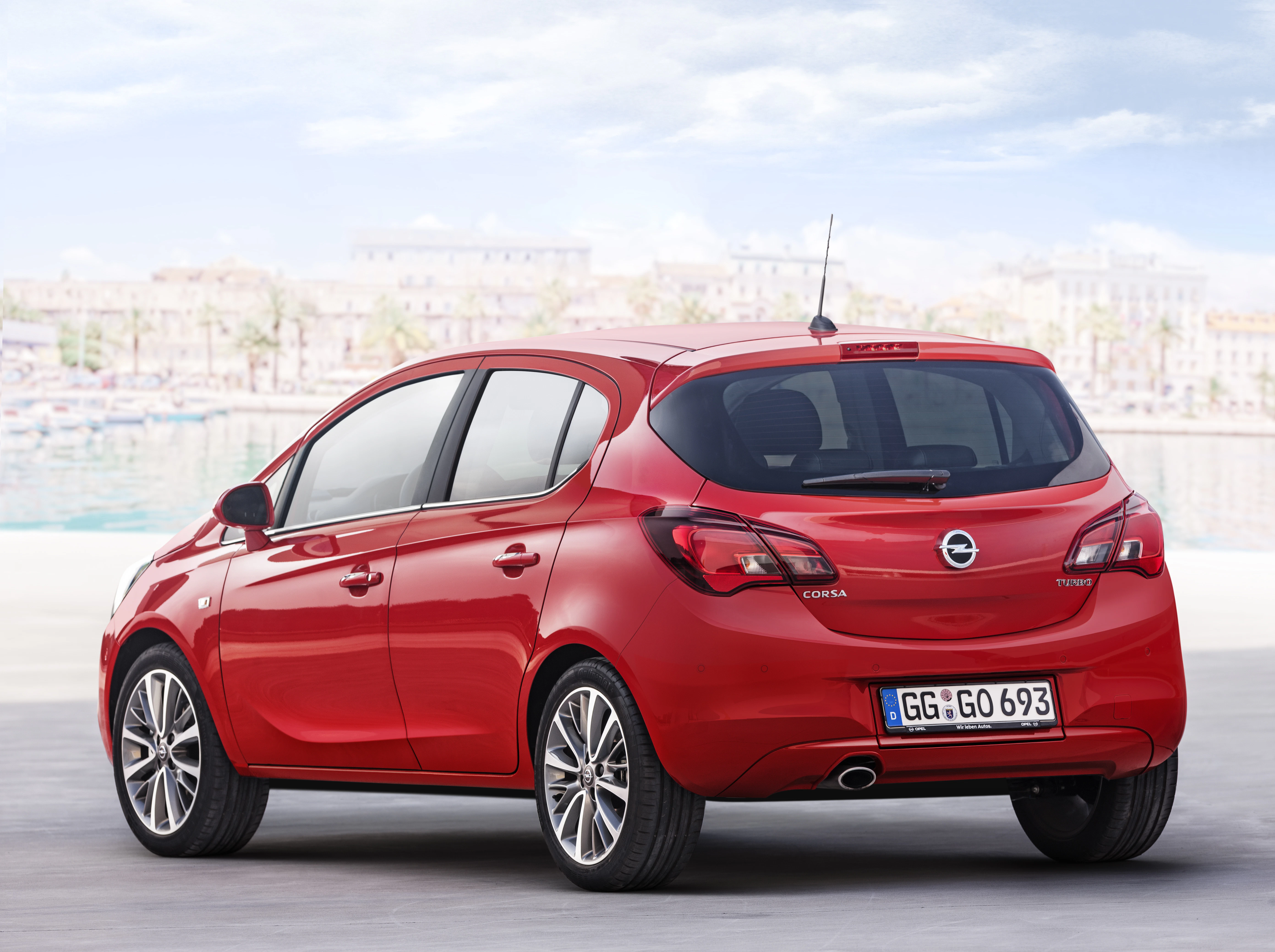 Roter Opel Corsa geparkt vor einer verschwommenen Stadtlandschaft. Ansicht von hinten, mit Dachantenne und großem Heckfenster.
