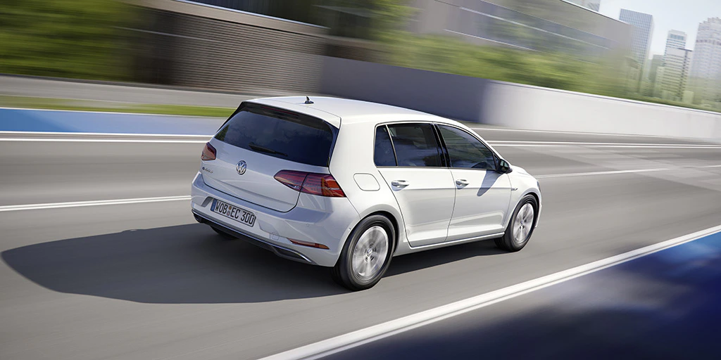 Weißer Volkswagen Golf fährt schnell auf einer Straße in städtischer Umgebung, Gebäude und Bäume im Hintergrund.