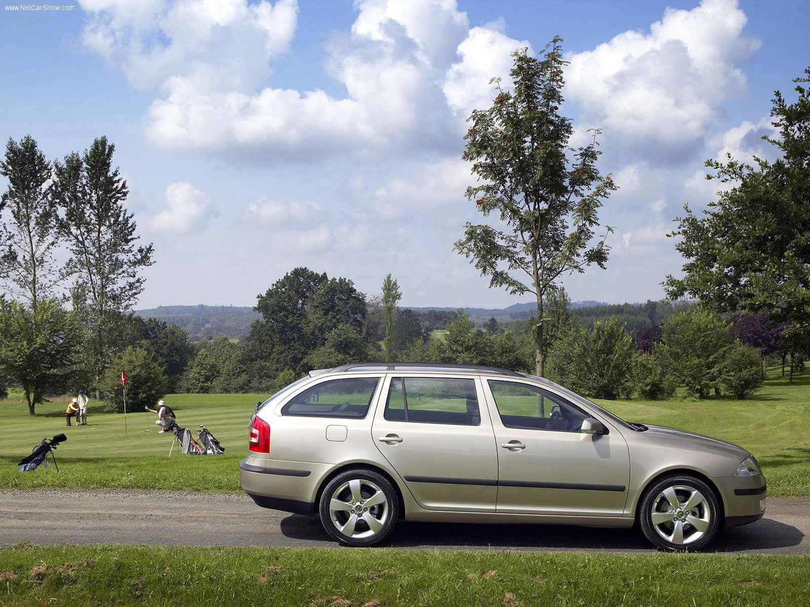 Silberner Kombi parkt nahe einem Golfplatz, auf dem Menschen Golf spielen. Bäume im Hintergrund unter einem blau bedeckten Himmel.