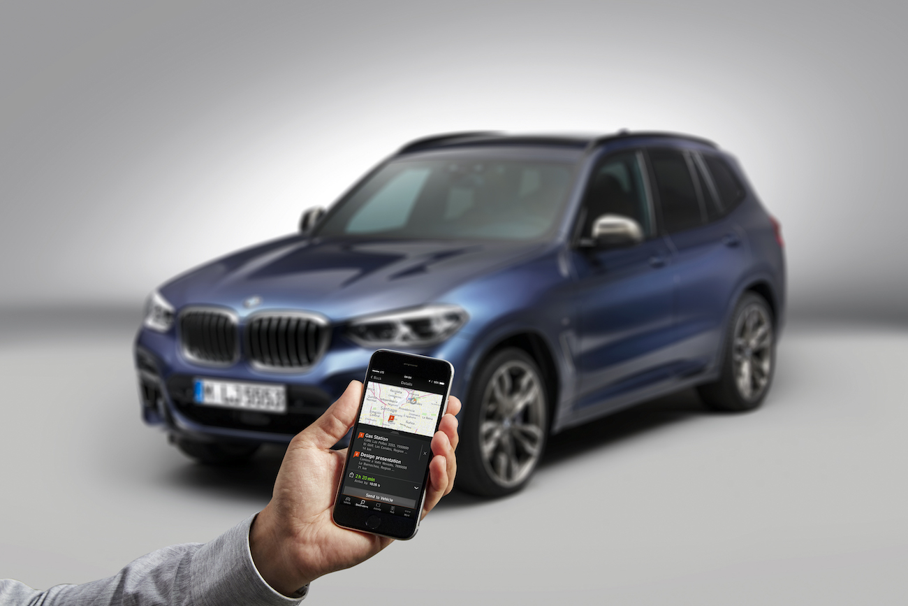 Eine Hand hält ein Smartphone mit Fahrzeug-App, im Hintergrund steht ein blauer SUV.