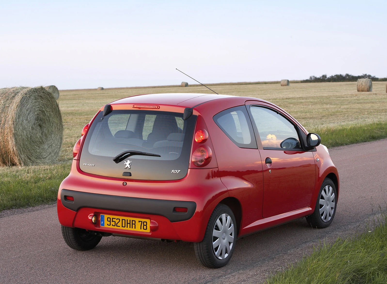 Roter Kleinwagen auf Landstraße, umgeben von grünen Feldern mit Heuballen im Hintergrund.