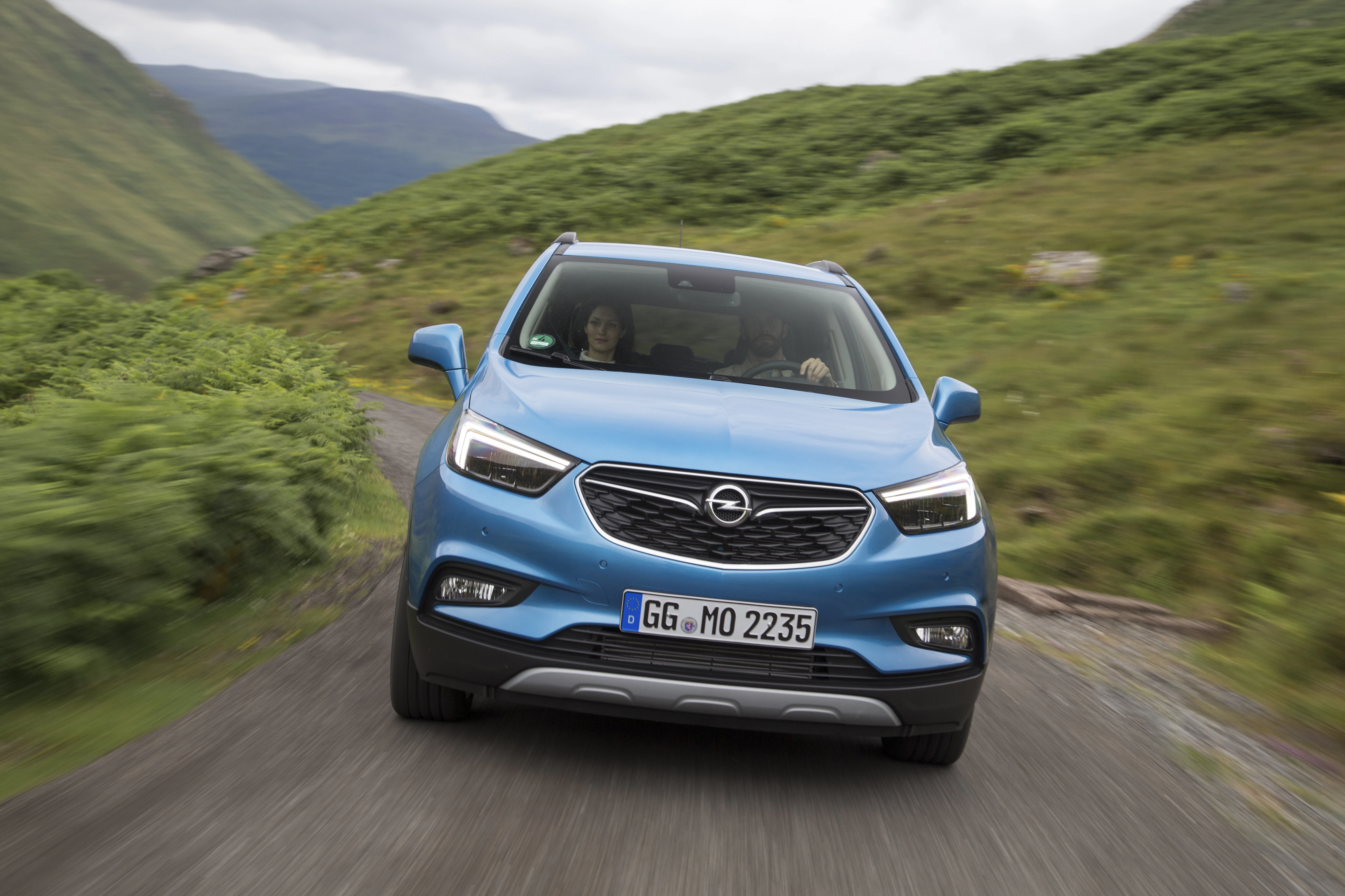 Blauer Opel SUV fährt auf schmaler Landstraße durch hügelige, grüne Landschaft. Fahrer und Beifahrer im Auto sichtbar.
